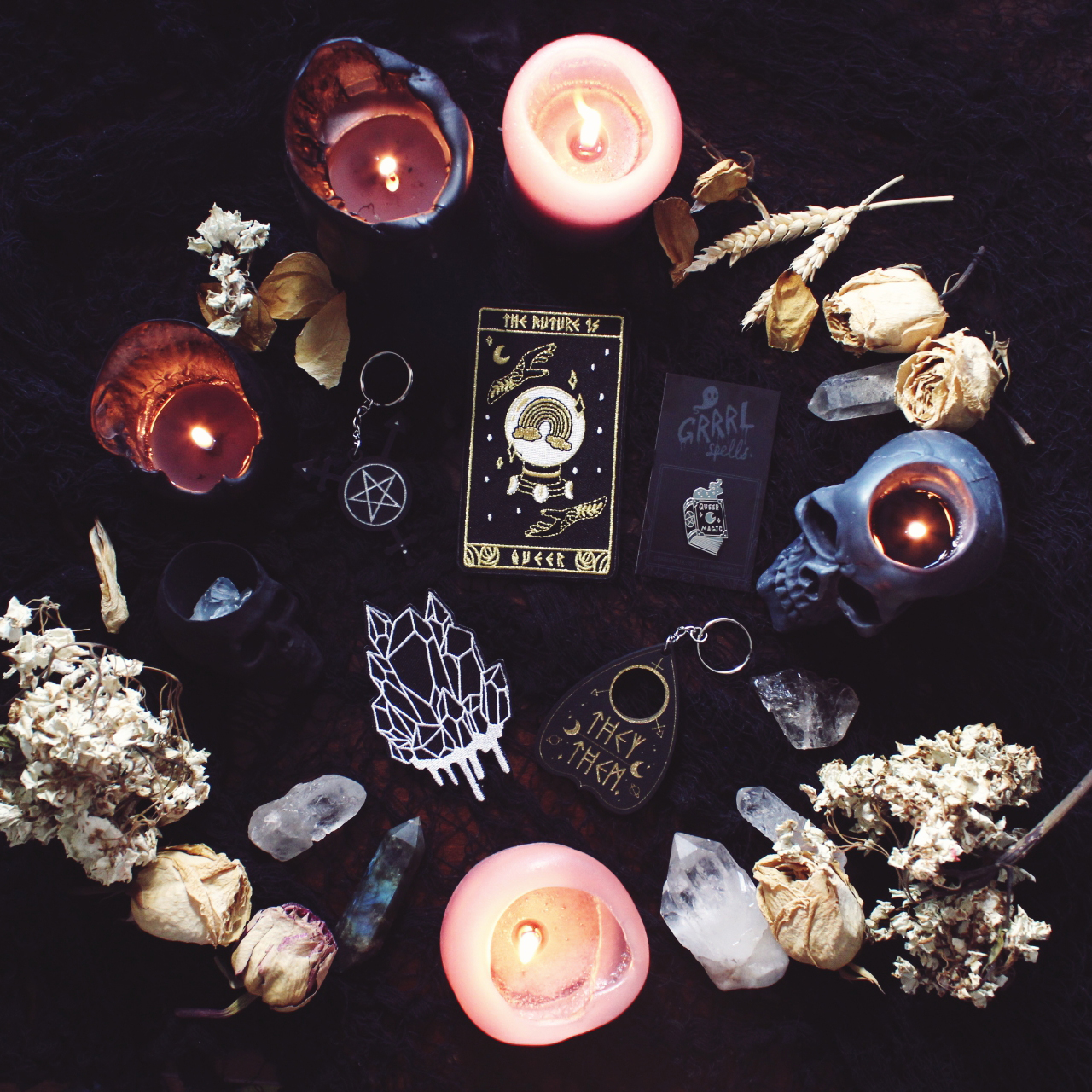 Image of crystals, dried flowers and lit candles surrounding crystal and tarot patches, and occult keychains and pins
