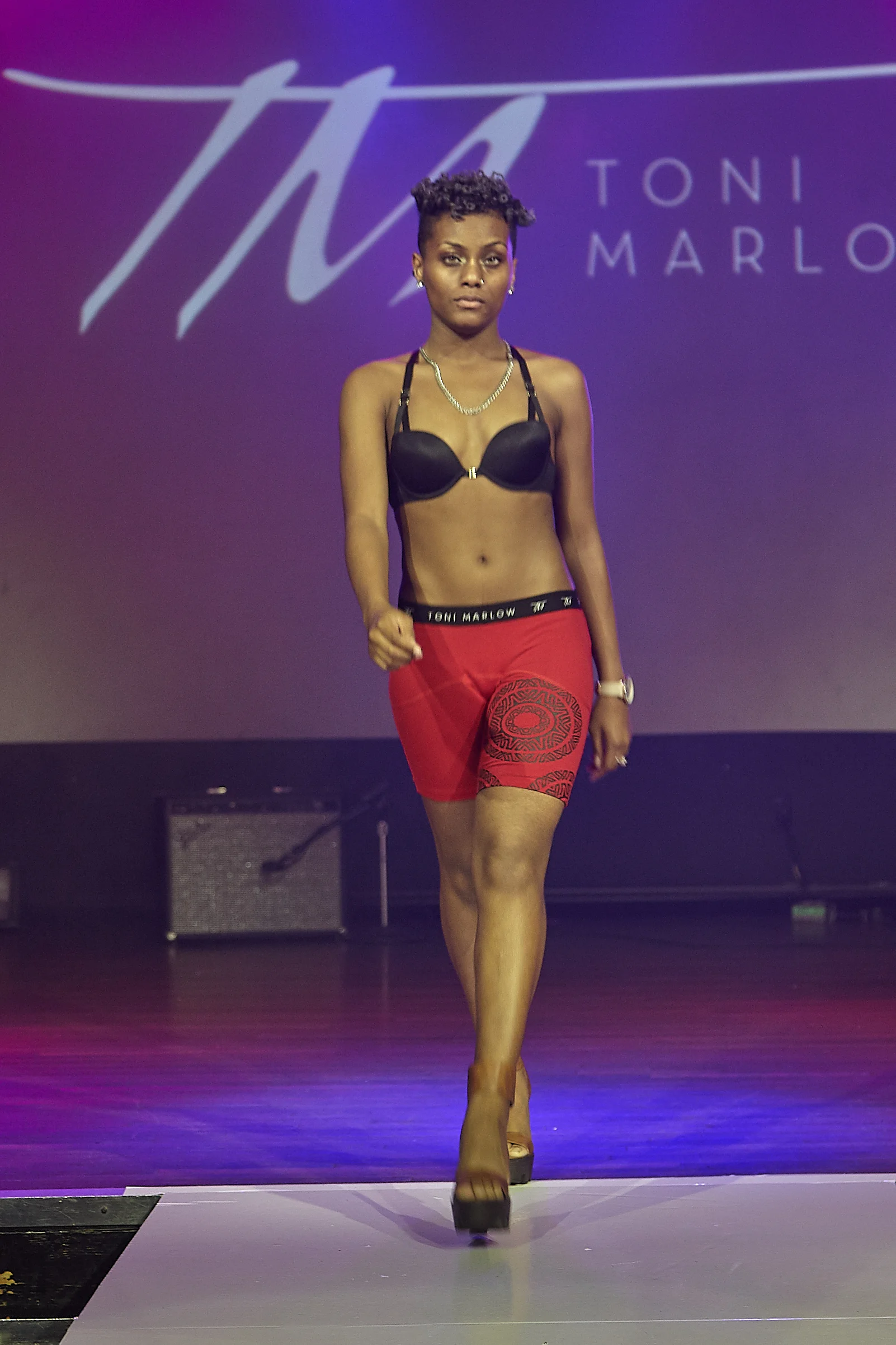 Image of model on runway in branded red boxer briefs, black bra, and gold neck chain