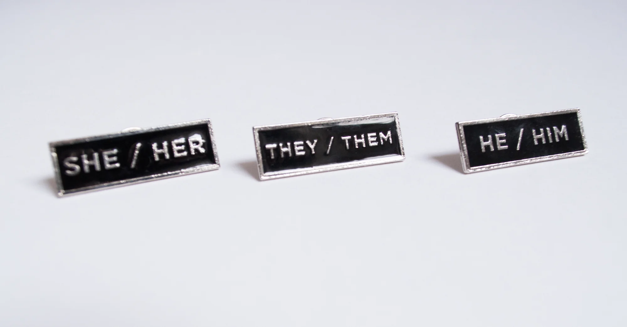 Image of three rectangular enamel pins with black background and pronouns in silver text (reading left to right), “she/her” “they/them” “he/him”