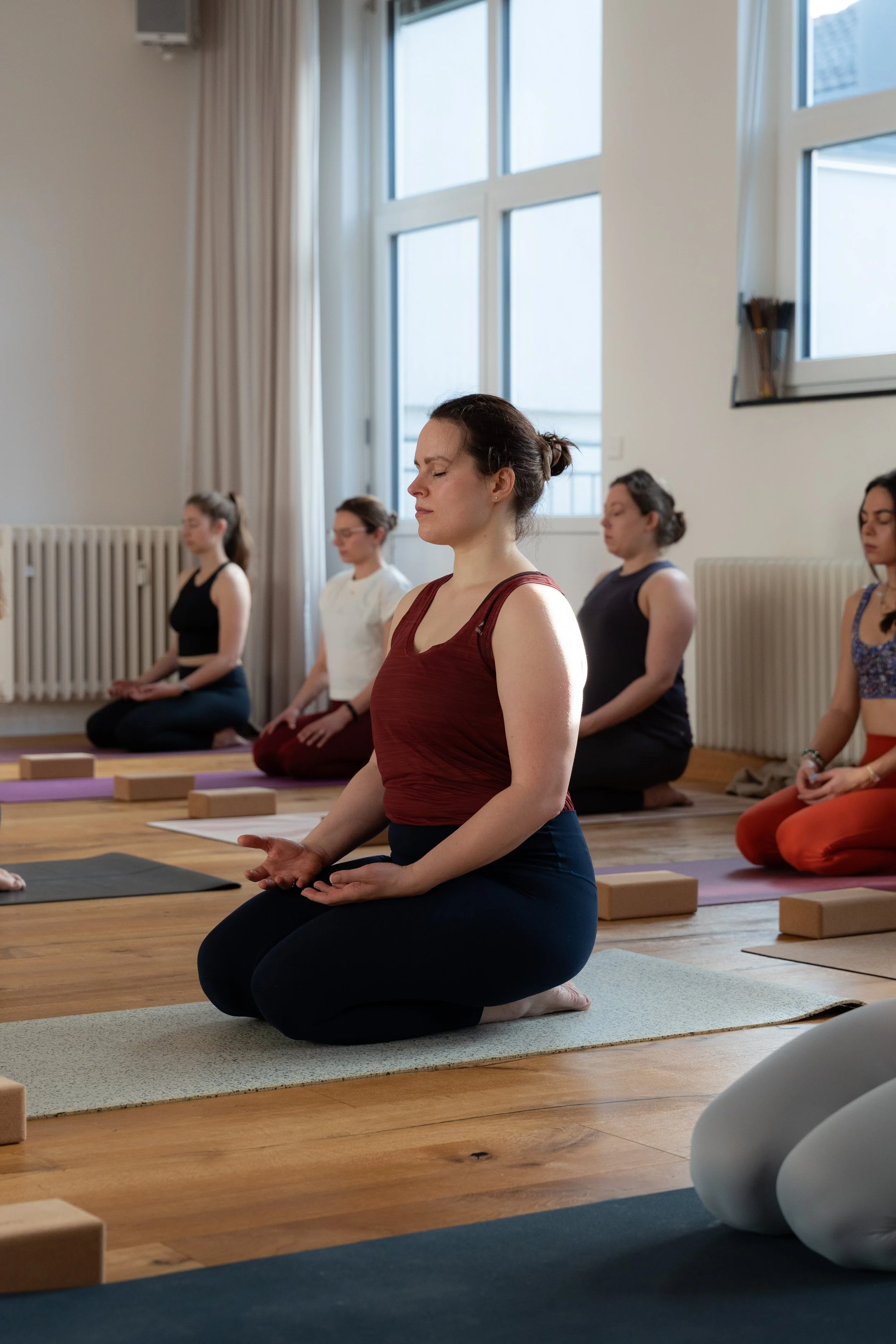 Eine Gruppe von Frauen sitzt in einer Yoga- oder Meditationsklasse auf Yogamatten in einem hellen Raum mit großen Fenstern, alle in Meditationshaltung mit geschlossenen Augen.