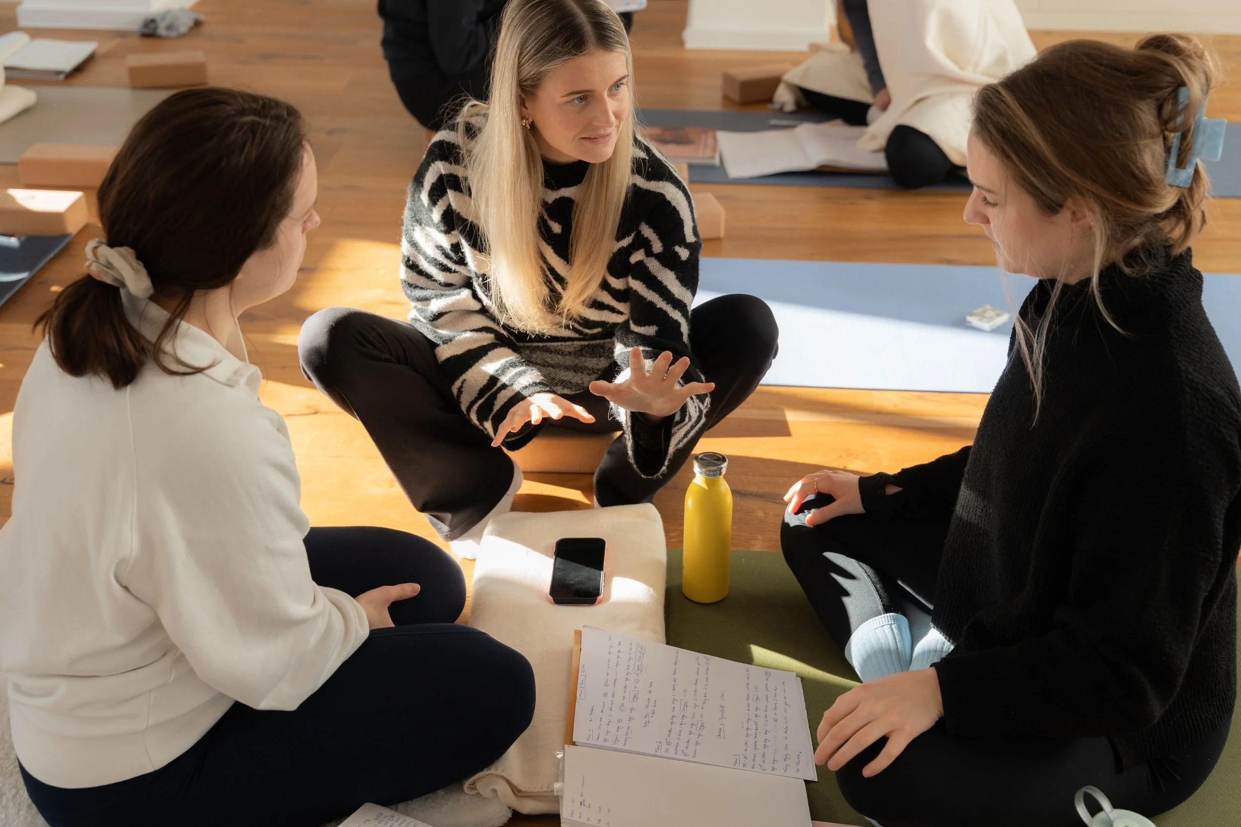 Drei Frauen sitzen auf einem Teppichboden, im Gespräch, umgeben von Notizbüchern, einem Smartphone und einer Wasserflasche.