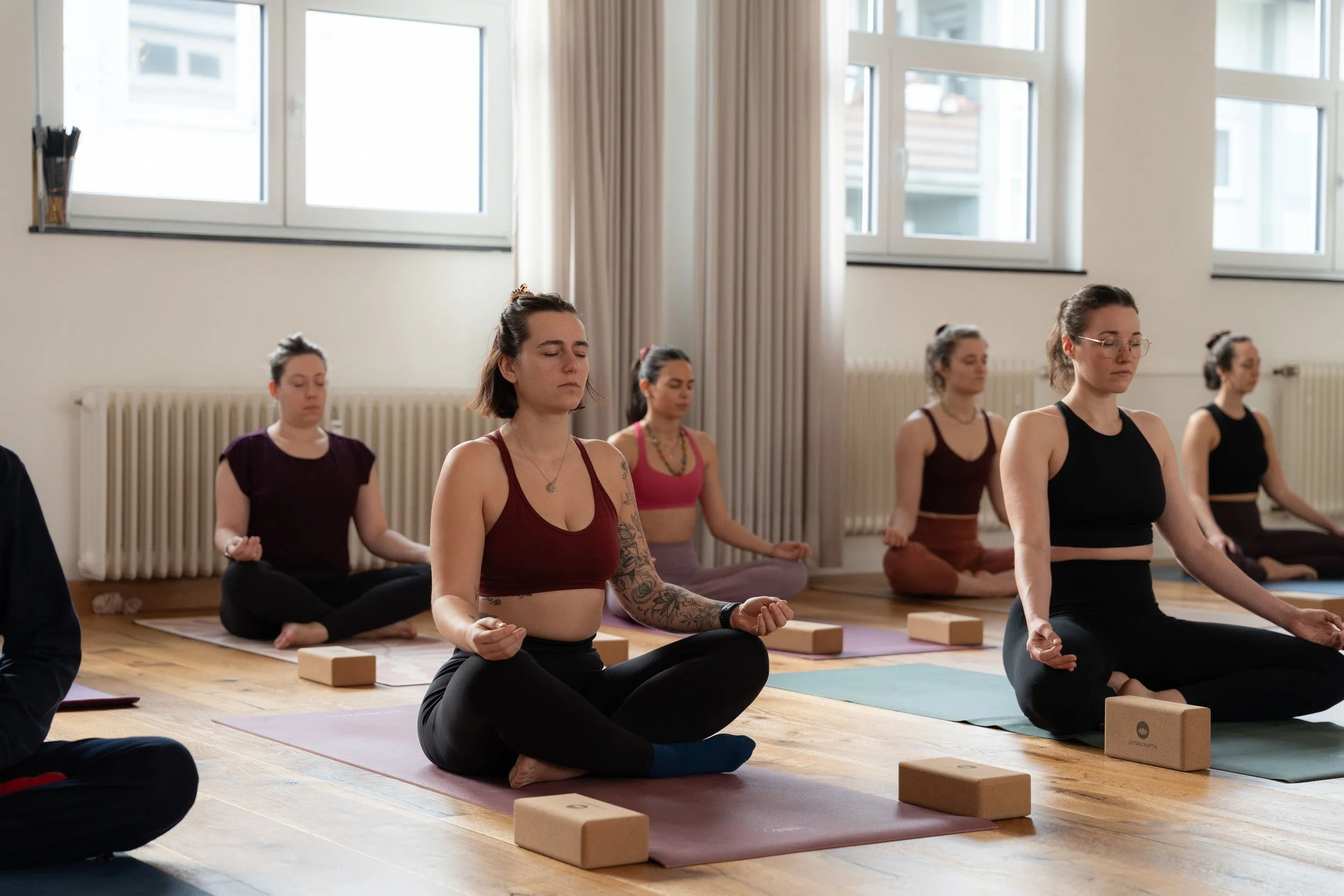Eine Gruppe von Frauen macht eine Yoga- oder Meditationssitzung in einem hellen Raum mit großen Fenstern.