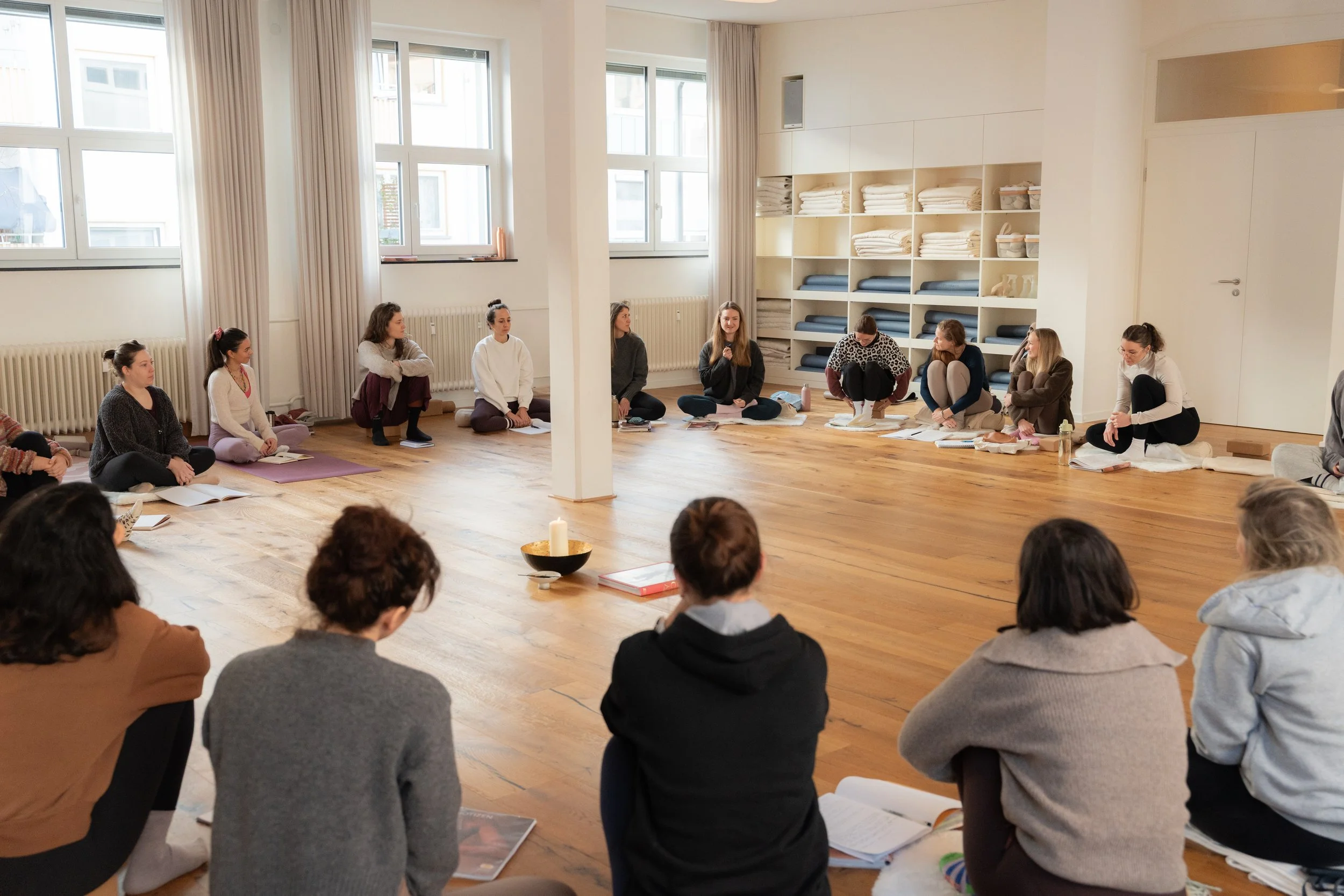 Eine Gruppe von Menschen sitzt in einem Kreis auf dem Boden in einem hellen Raum mit großen Fenstern, auf einem Teppich, mit einer ruhigen Atmosphäre.