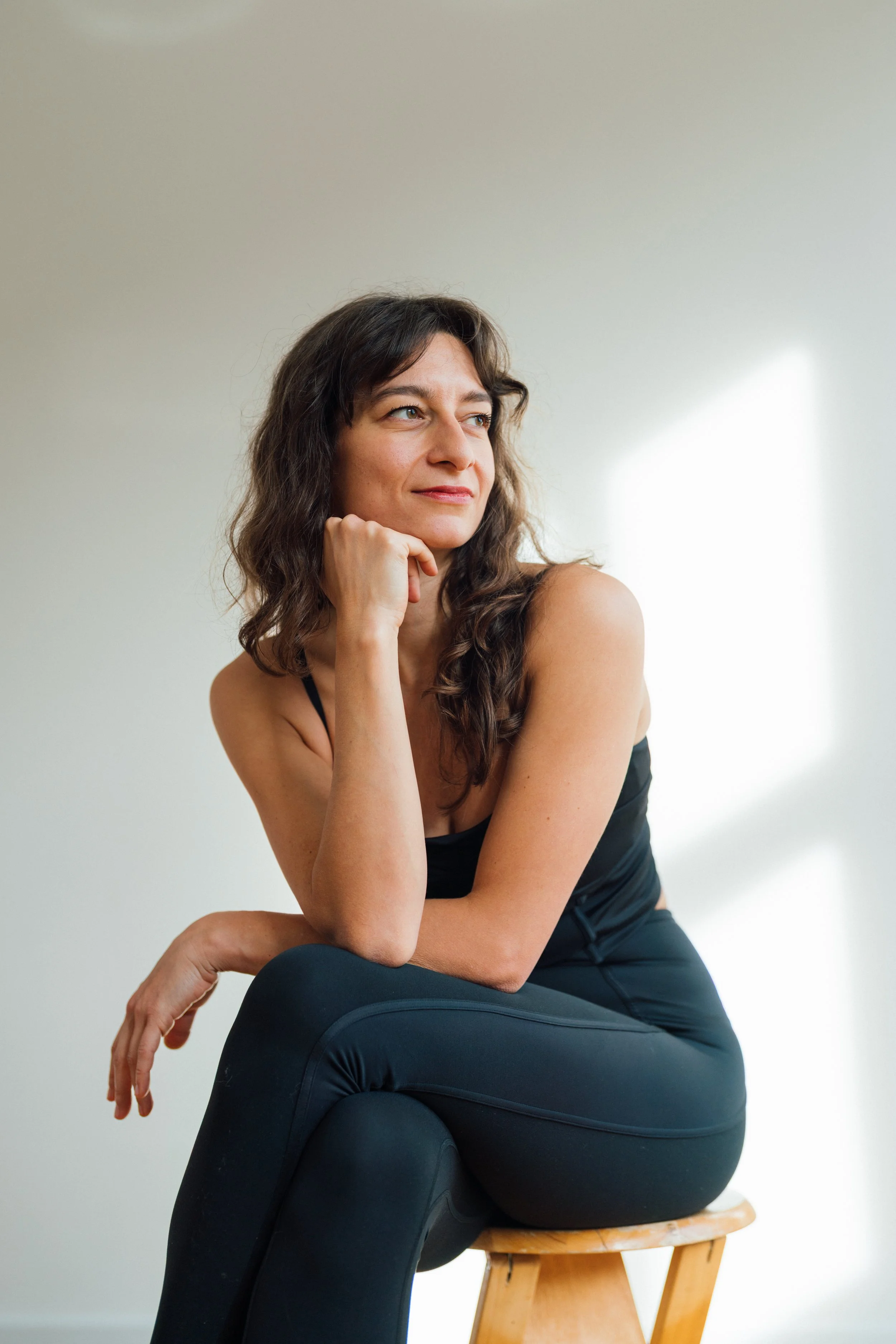 Frau sitzt nachdenklich auf einem Holzstuhl, trägt schwarze Sportkleidung, mit Sonnenlicht im Hintergrund an einer weißen Wand.