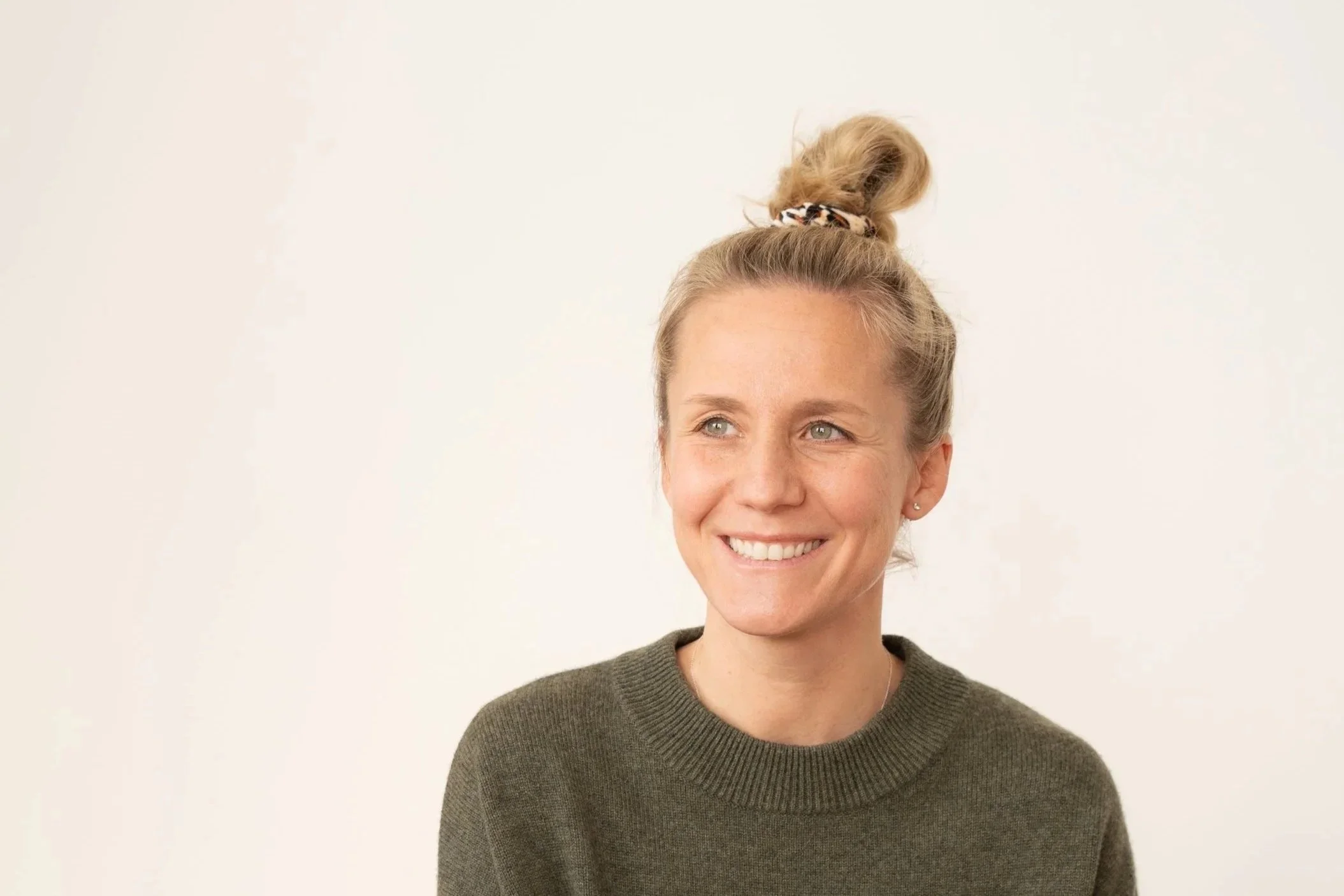 Junge Frau mit blonden Haaren in einem Dutt, lächelnd, gegen weißen Hintergrund, trägt grauen Pullover.