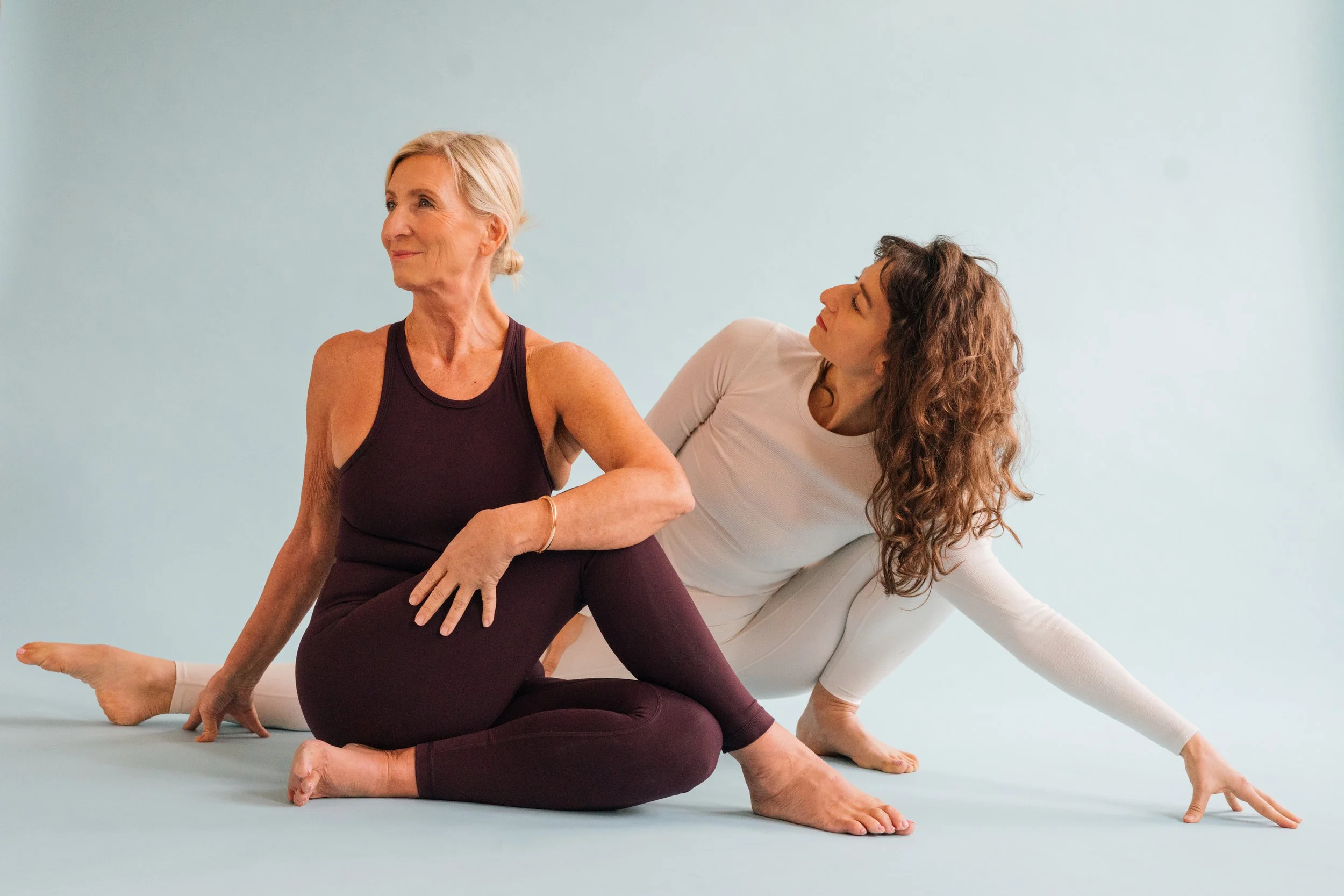 Zwei Frauen üben Yoga auf einer hellblauen Matte, eine ältere Frau sitzt in einer halb gekreuzten Haltung, die jüngere Frau hilft ihr, eine Dehneposition einzunehmen.