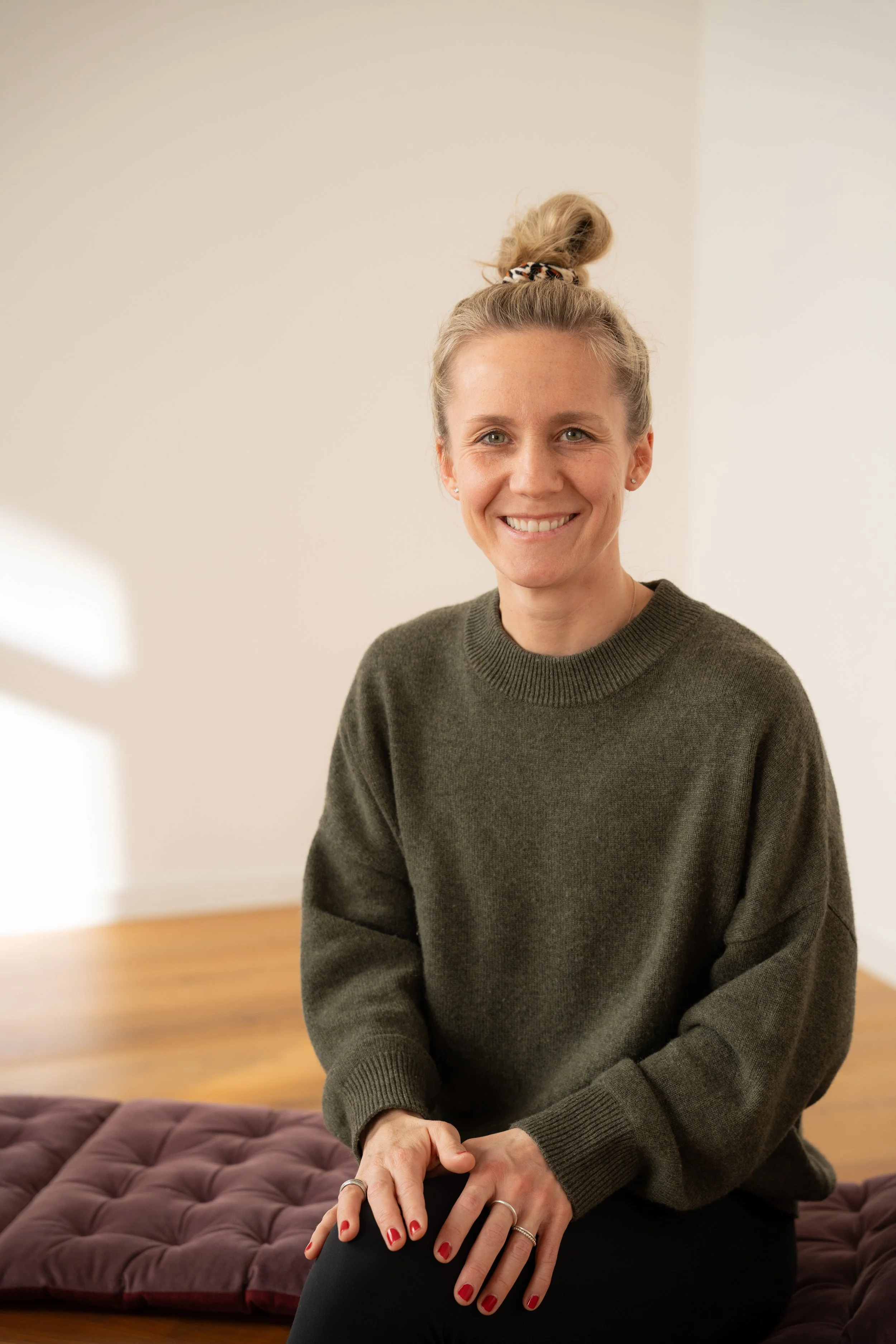 Eine lächelnde Frau mit blonden Haaren in einem hohen Dutt sitzt auf einer gepolsterten Fläche in einem Raum mit weißen Wänden und Holzboden.