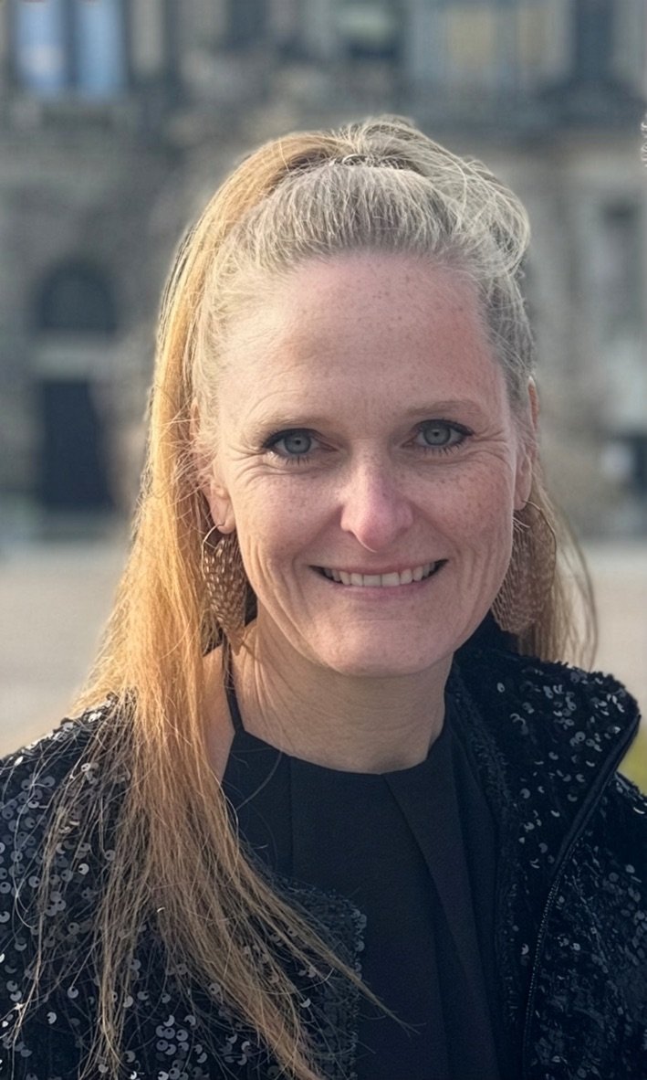 Lächelnde Frau mit roten Haaren, die einen Pferdeschwanz trägt, vor einer urbanen Kulisse bei Sonnenuntergang.