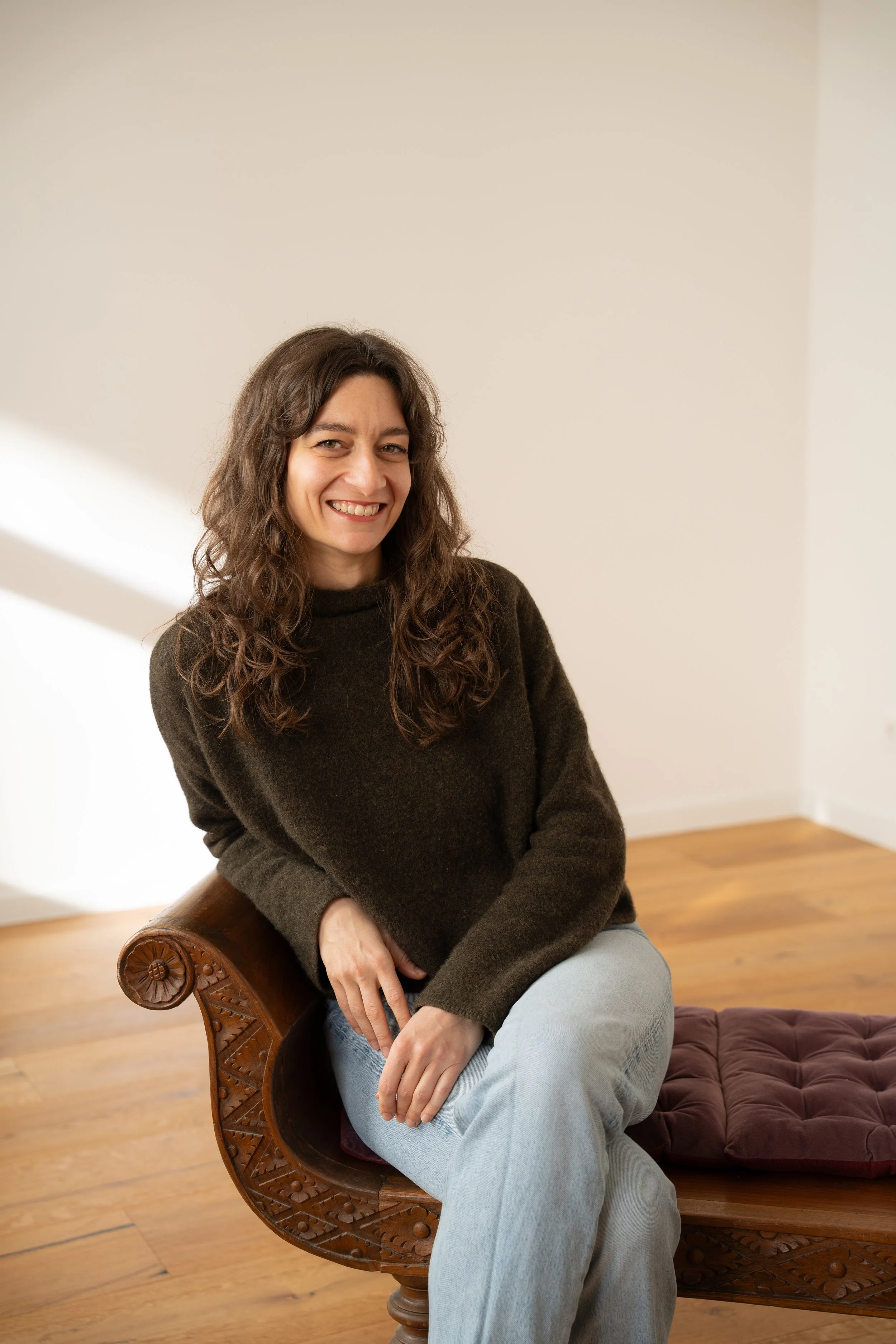 Eine lächelnde Frau mit braunen, gewellten Haaren sitzt auf einer Holzbank in einem Raum mit Holzboden. Sie trägt einen braunen Pullover und helle Jeans.