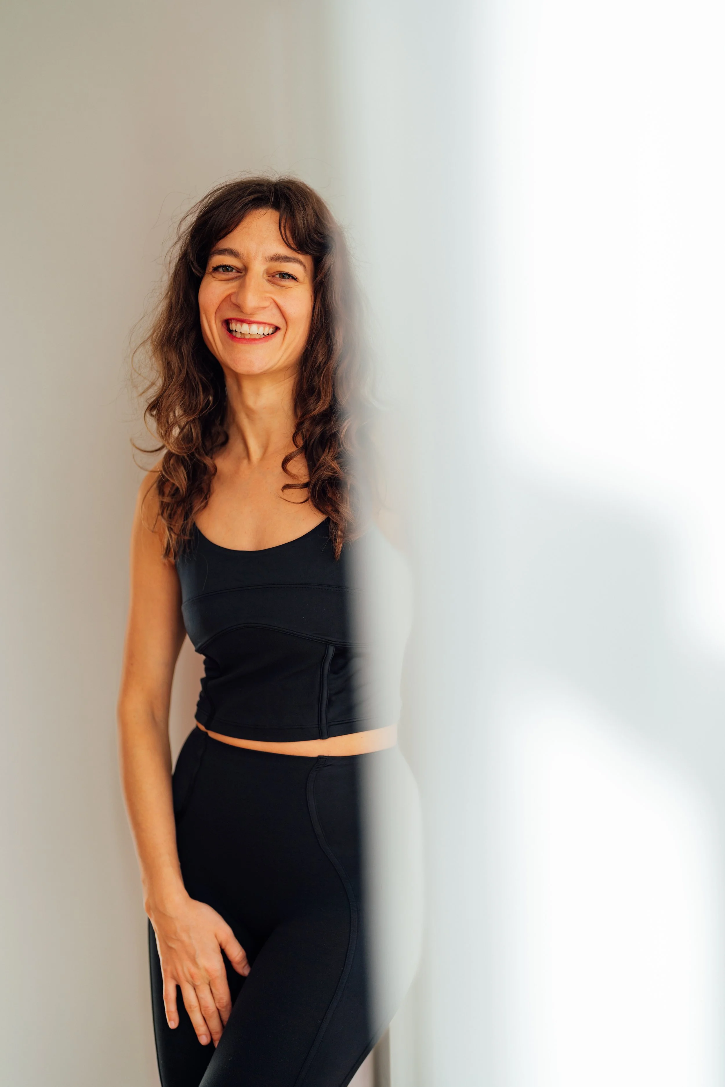 Lächelnde Frau in schwarzem Sportoutfit, halb hinter einer weißen Wand versteckt.