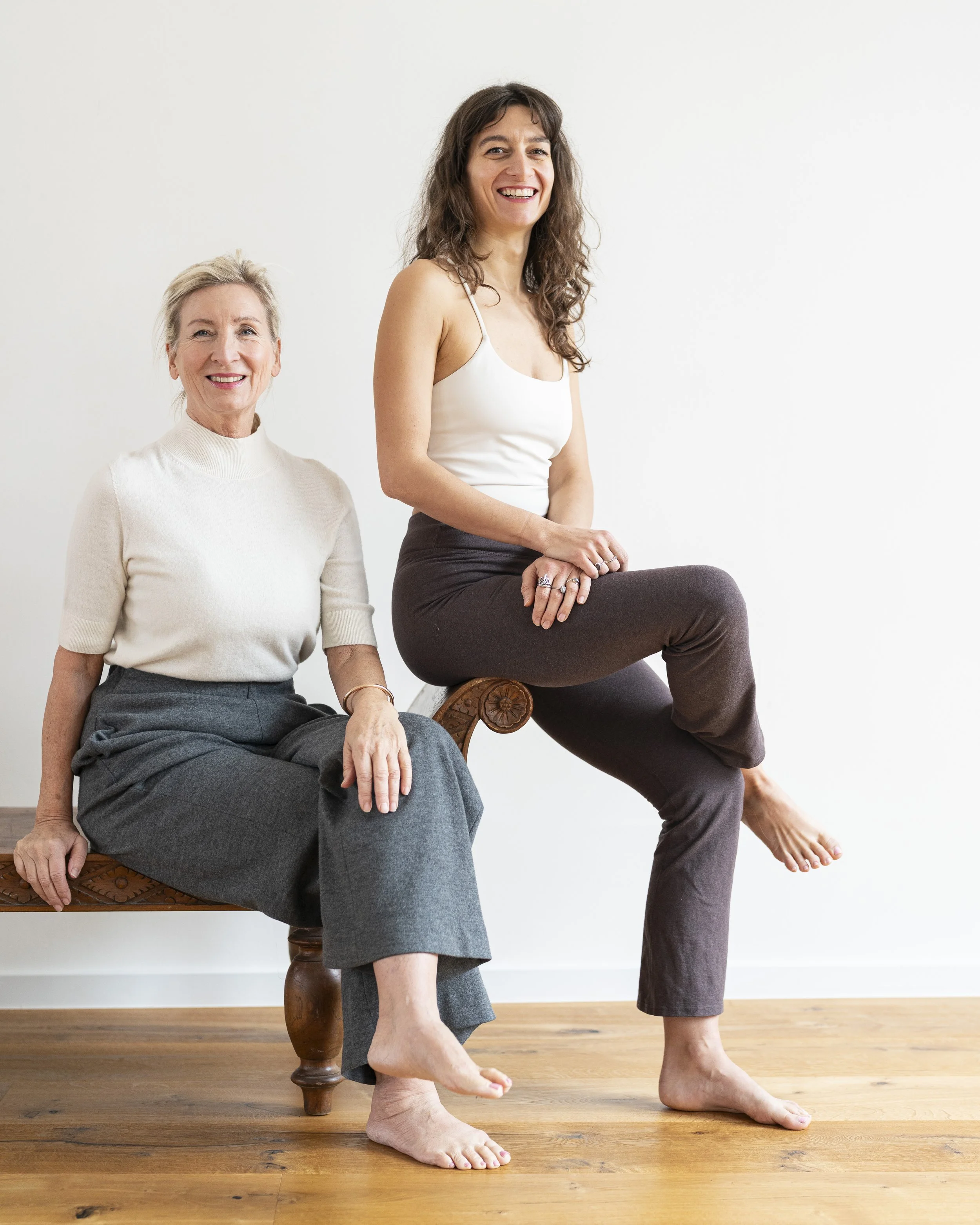 Zwei Frauen sitzen auf einer antiken Holzbank vor einer weißen Wand, lächelnd, barfuß, im modernen, eleganten Kleidungsstil.