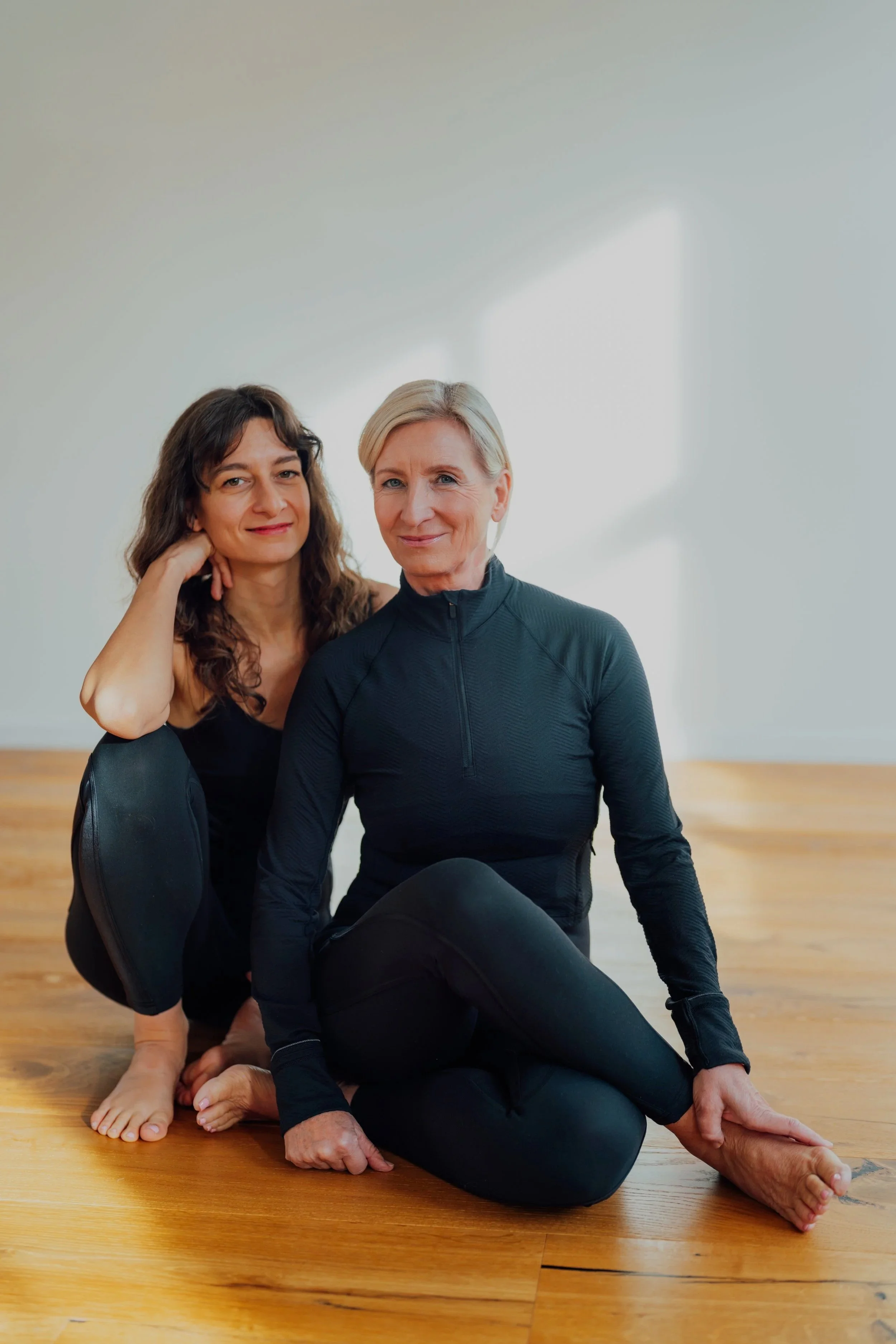 Zwei Frauen in Sportkleidung beim Yoga auf einem Holzfußboden in einem hellen Raum.