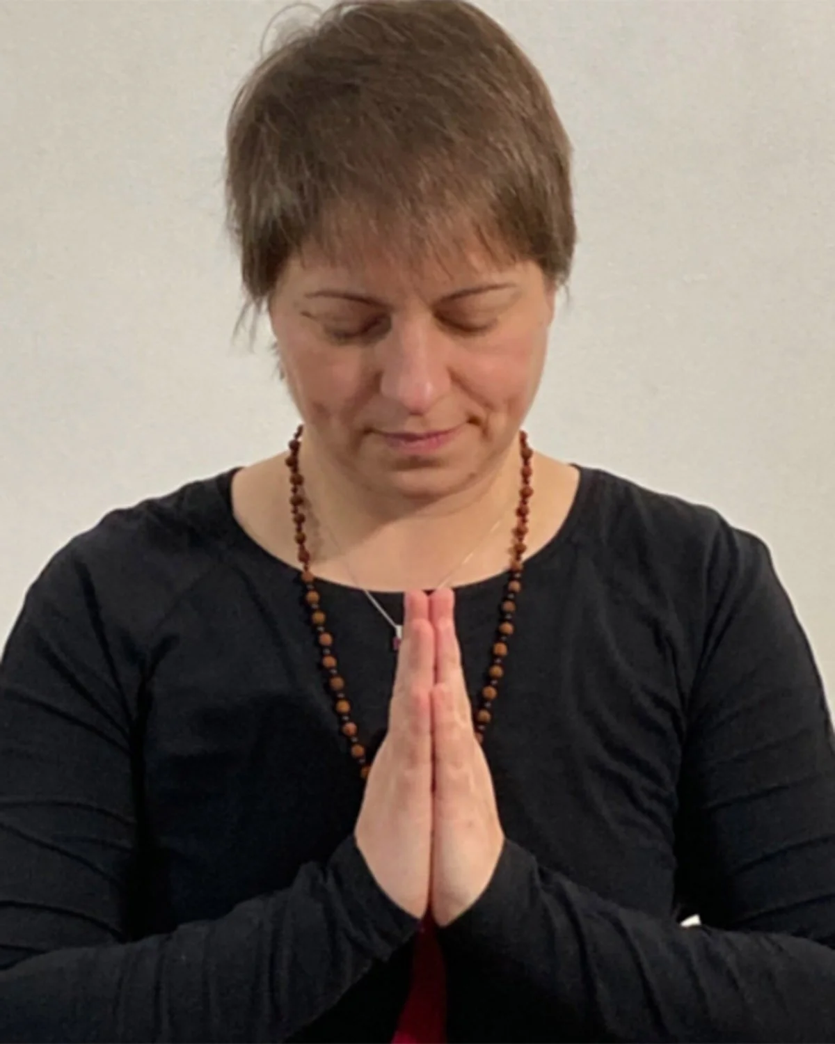 Frau mit kurzen braunen Haaren in Gebetshaltung, Hände vor der Brust, trägt schwarze Kleidung, einen braunen Gebetsschnur und eine kleine Kette, steht vor weißem Hintergrund.