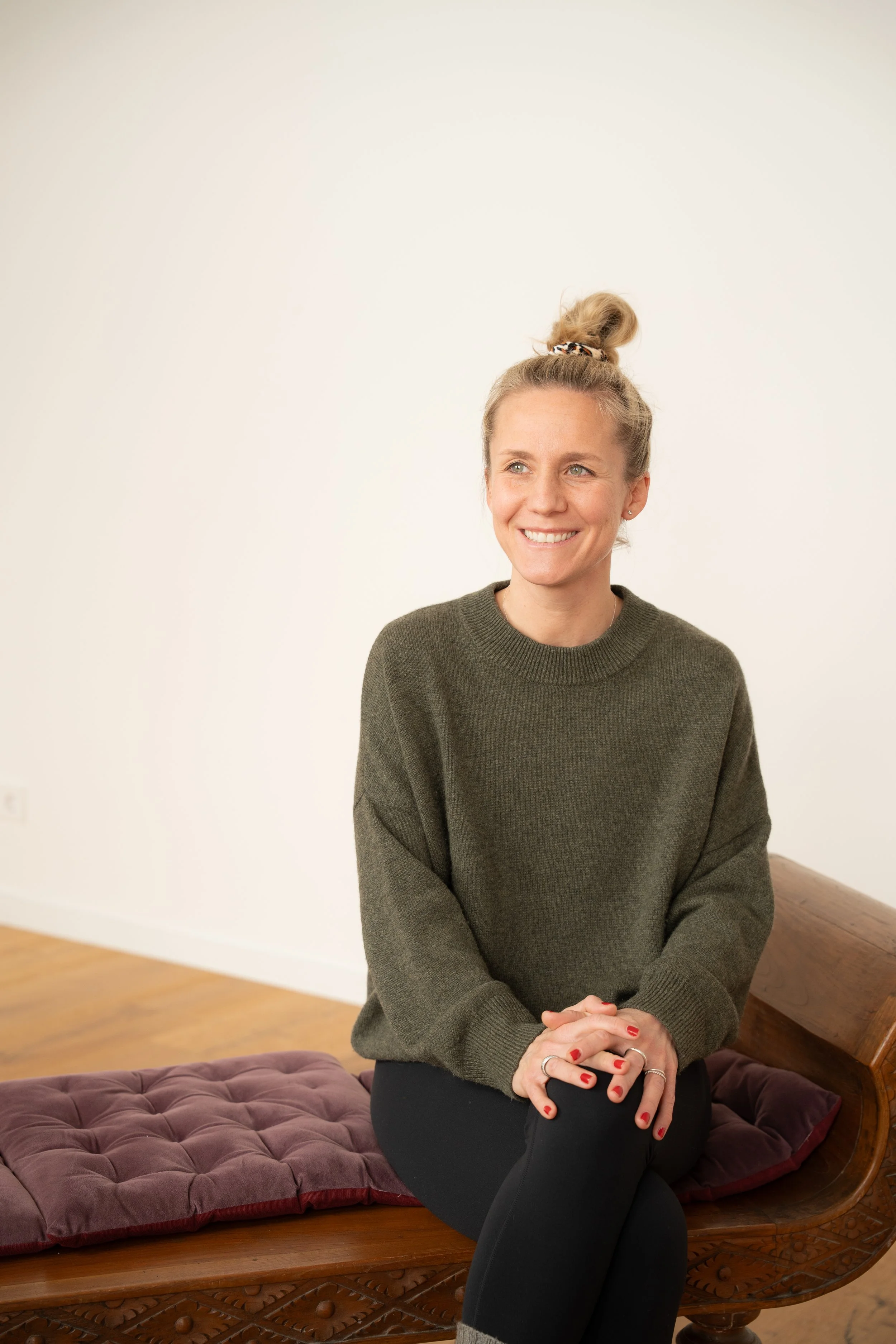 Junge Frau mit Blasshaut, blonden Haaren im Dutt, lächelnd, sitzend auf einer alten Holzbank mit rotem Polster, in einem Raum mit weißem Wandhintergrund.