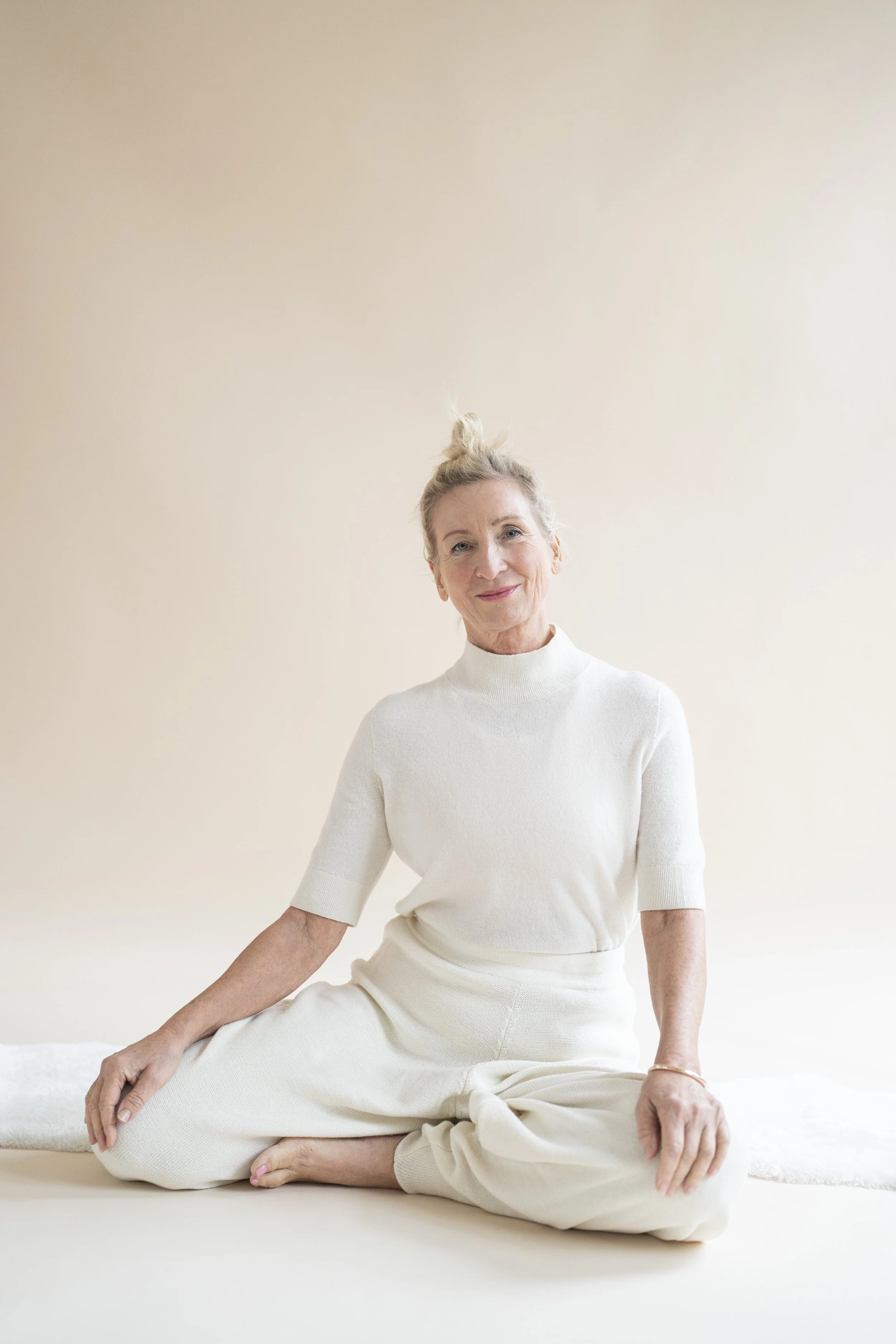 Ältere Frau in weißen Freizeitkleidern sitzt auf einem weißen Boden, vor einem neutralen Hintergrund, lächelnd in die Kamera schauend.