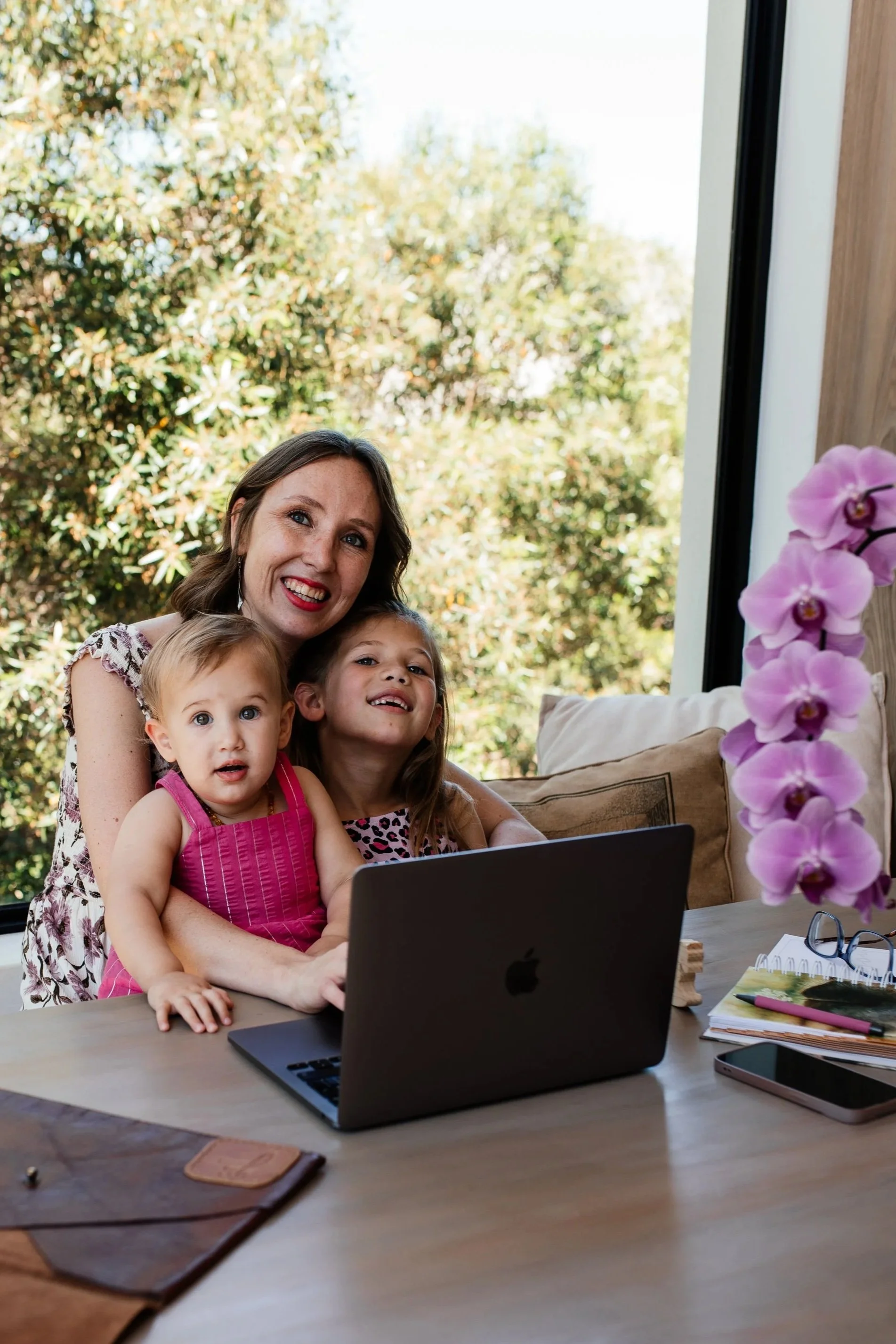 A Social media manger & mom of two young girls sitting together at a table with a laptop, smiling, with a window and greenery outside in the background.