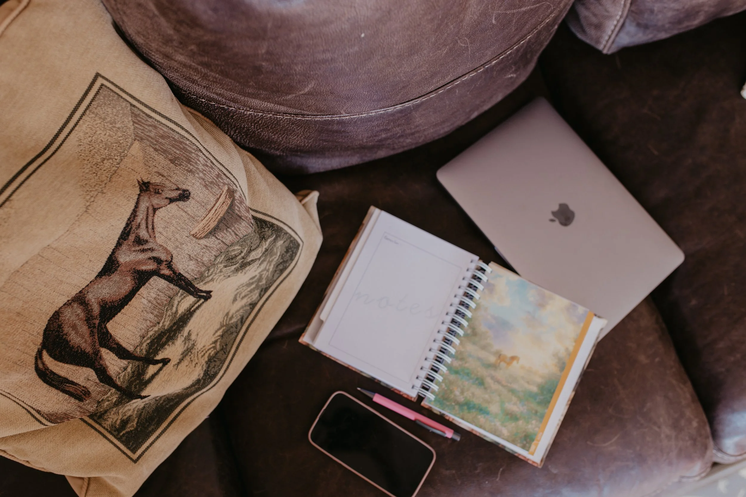 A cluttered brown leather couch with a decorative pillow featuring a running horse and a barn scene, an open spiral CM Paper Collective weekly Planner and a closed silver MacBook laptop, a pink pen, and a pink and black smartphone.