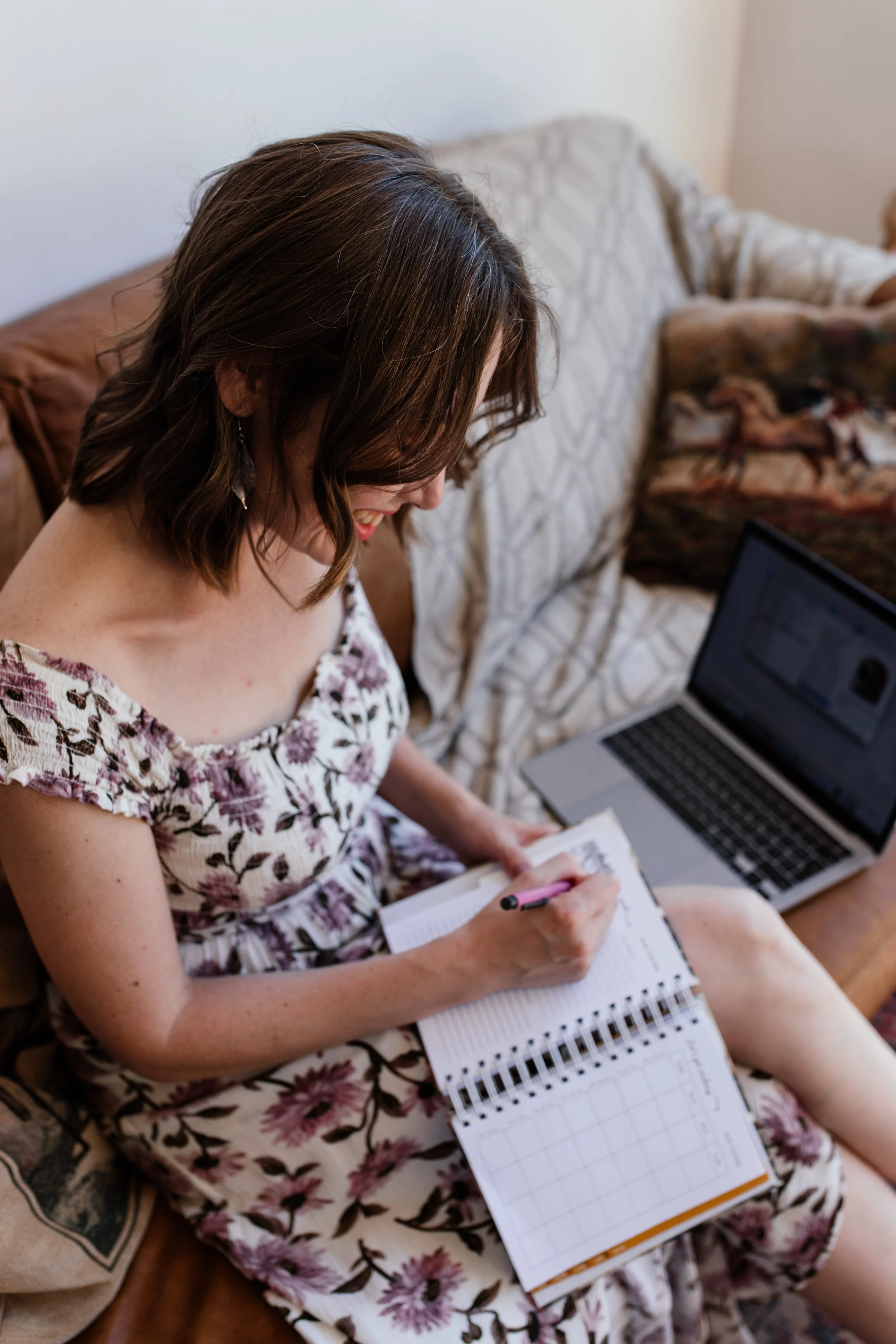 Social media manger sitting on a couch, smiling and writing in a planner with a pen, with a laptop on her lap, in a cozy living room.
