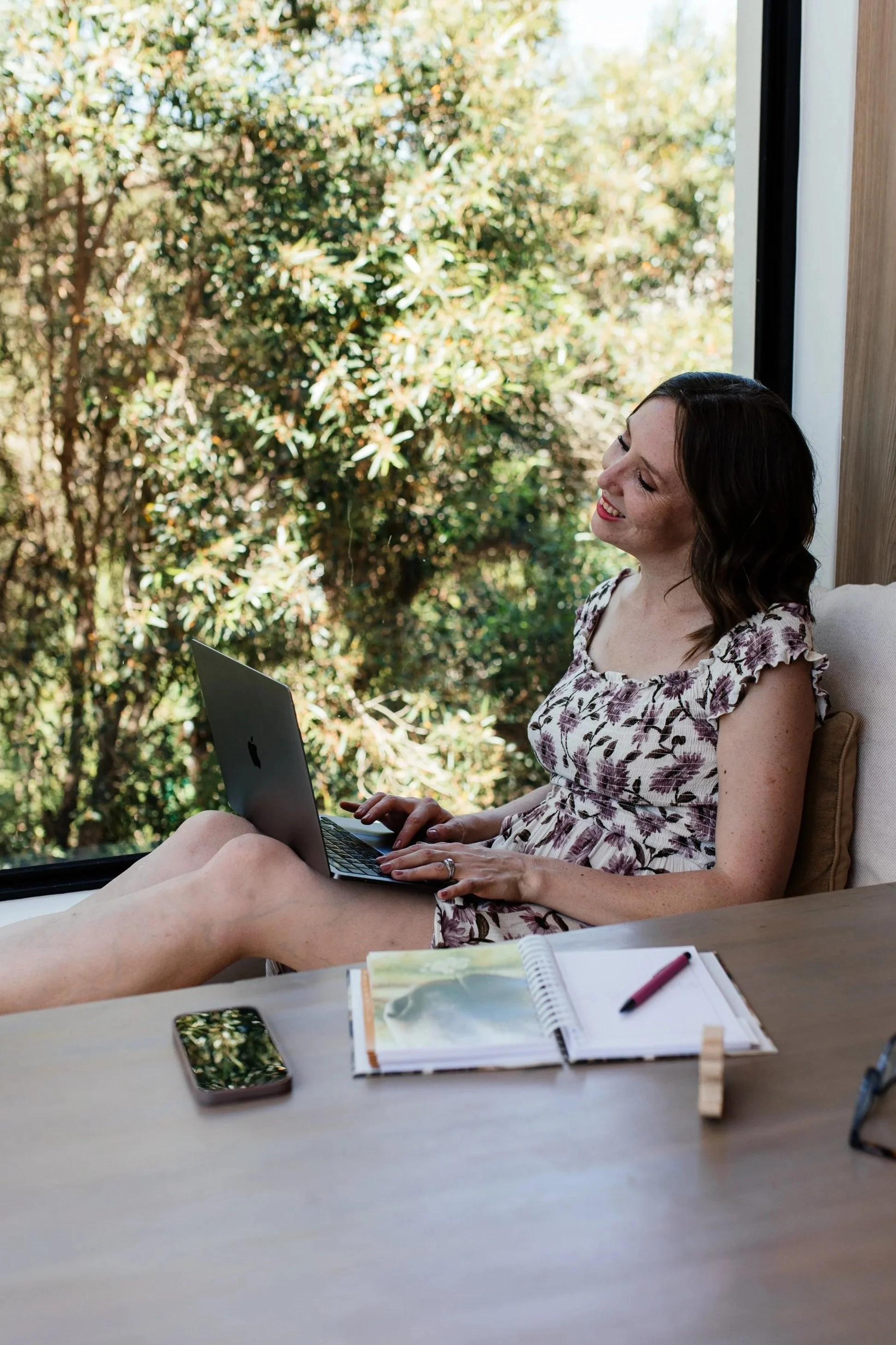 Social media manger sitting by a large window with trees outside, working on a laptop, with notebooks, a pen, a smartphone, and glasses on a wooden table.