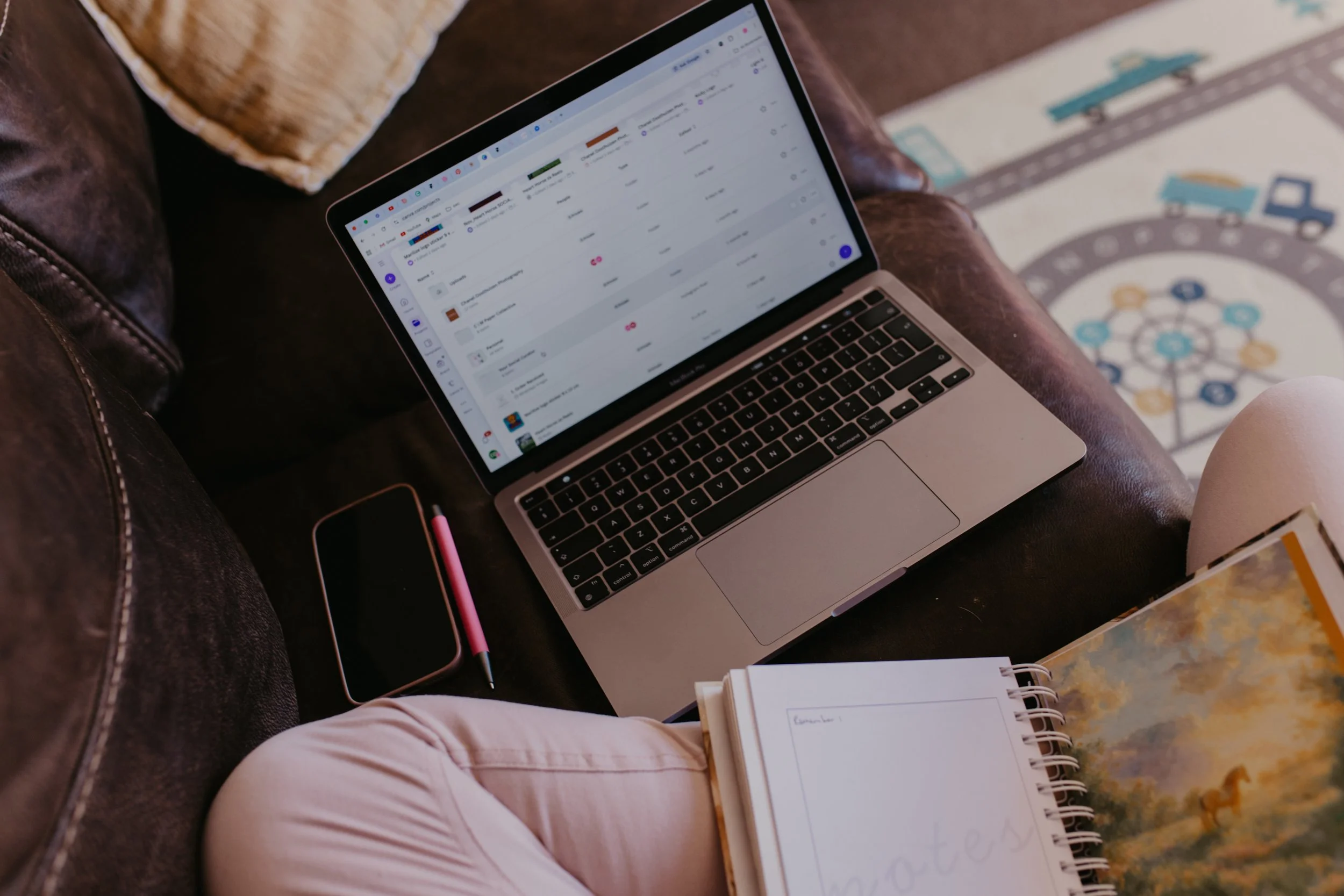 A Social media Manager sitting on a brown leather couch working on a laptop with a pink pen and a smartphone nearby, and a notebook and a children's book on the person's lap. There is a colorful rug with a wheel and truck design on the floor.