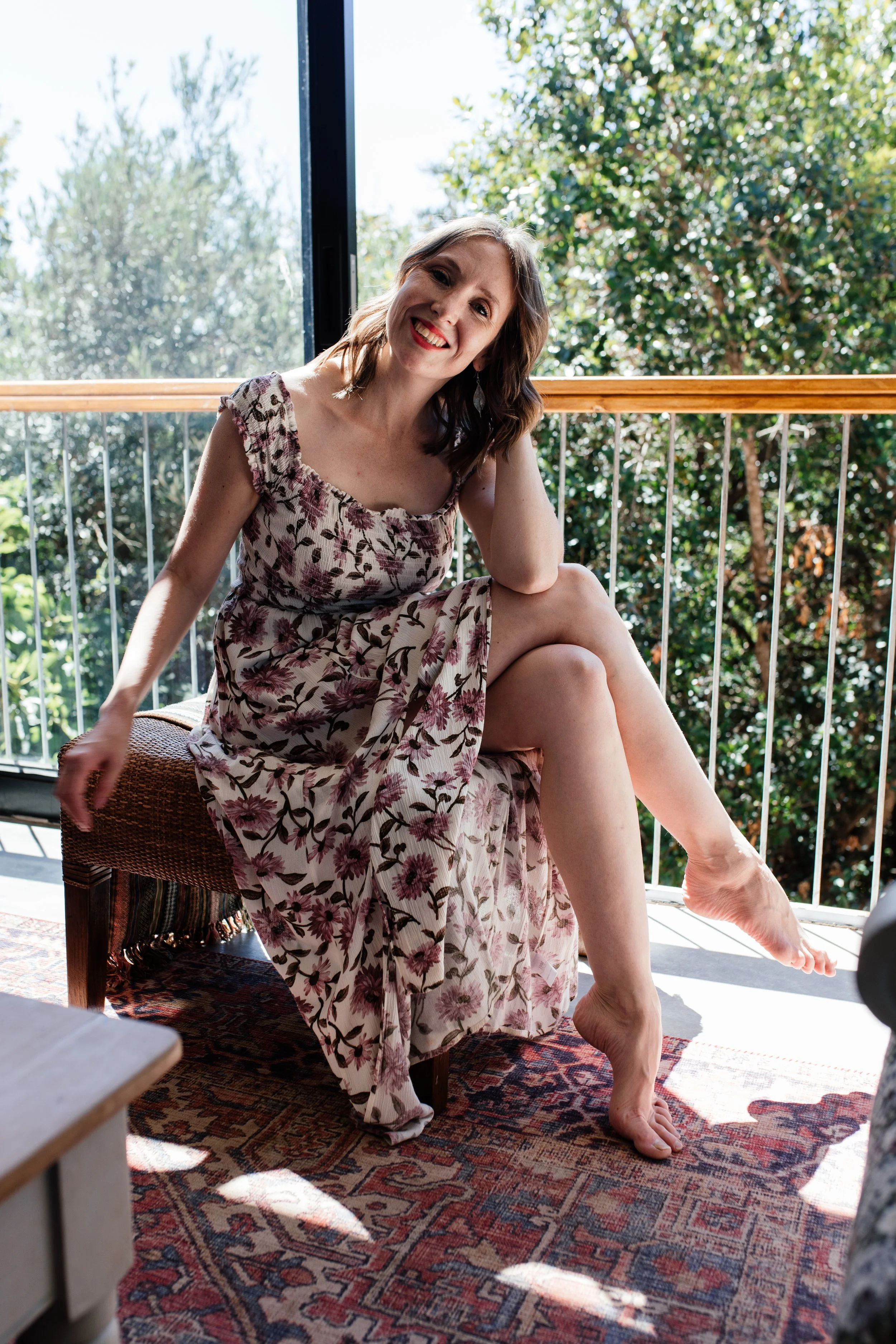 A Social media manger in a floral dress sitting on a cushion on a balcony, smiling at the camera with greenery outside.