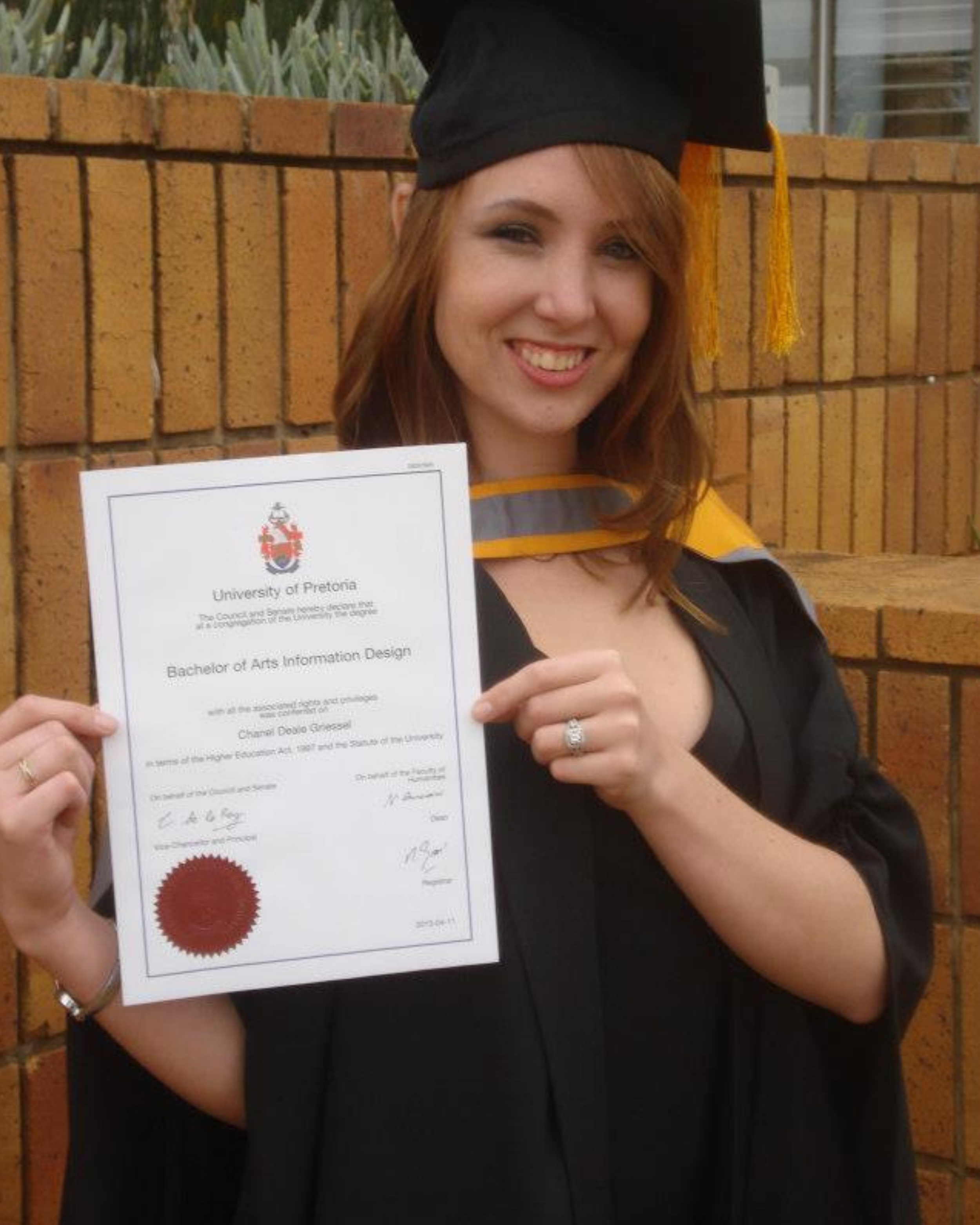 A woman in graduation attire holding a diploma in front of a brick wall, smiling at the camera.