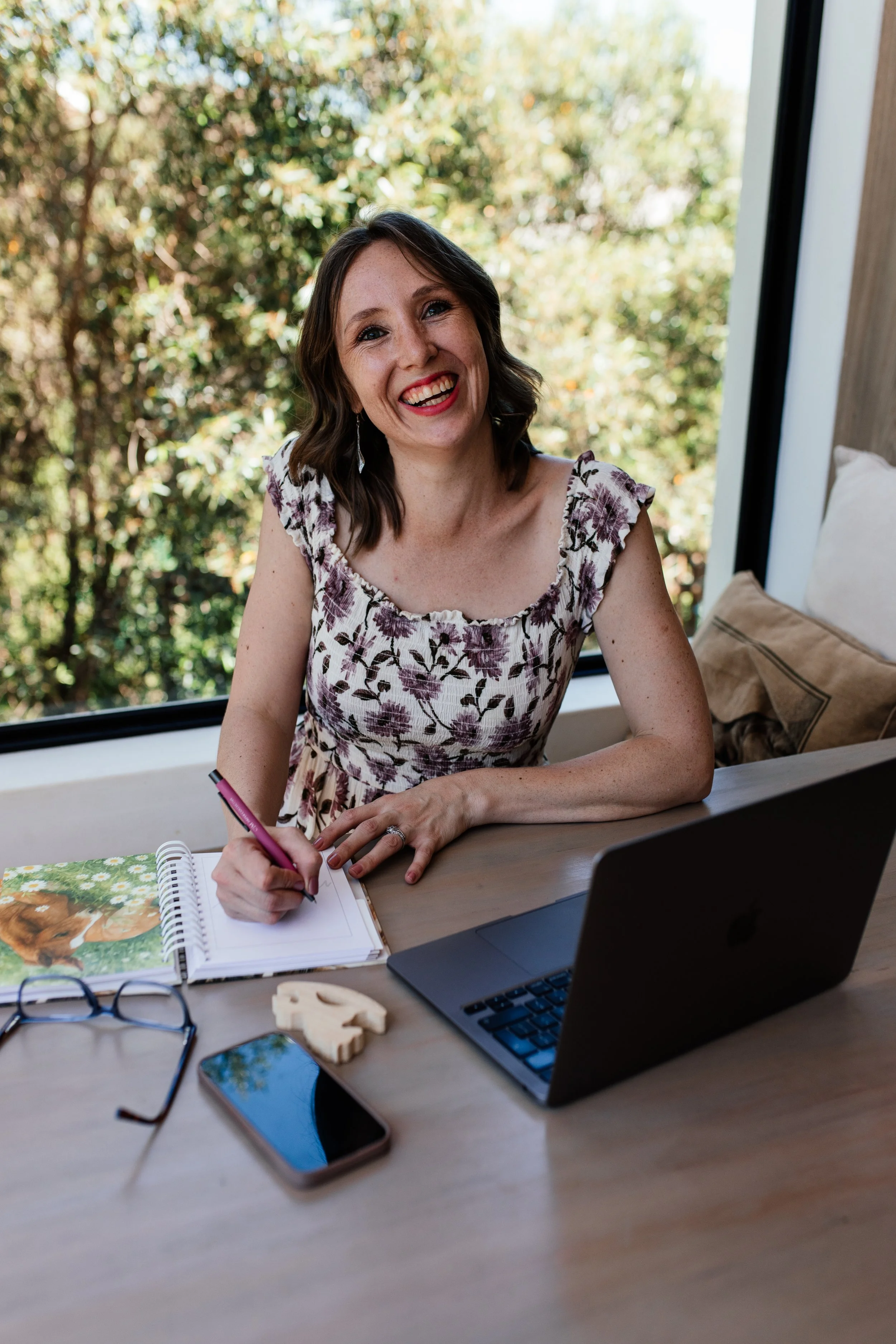 A Social Media manager smiling, holding a pink pen, with a notebook in front of her. On the table are a pair of glasses, a smartphone, a small decorative object, and a closed laptop.