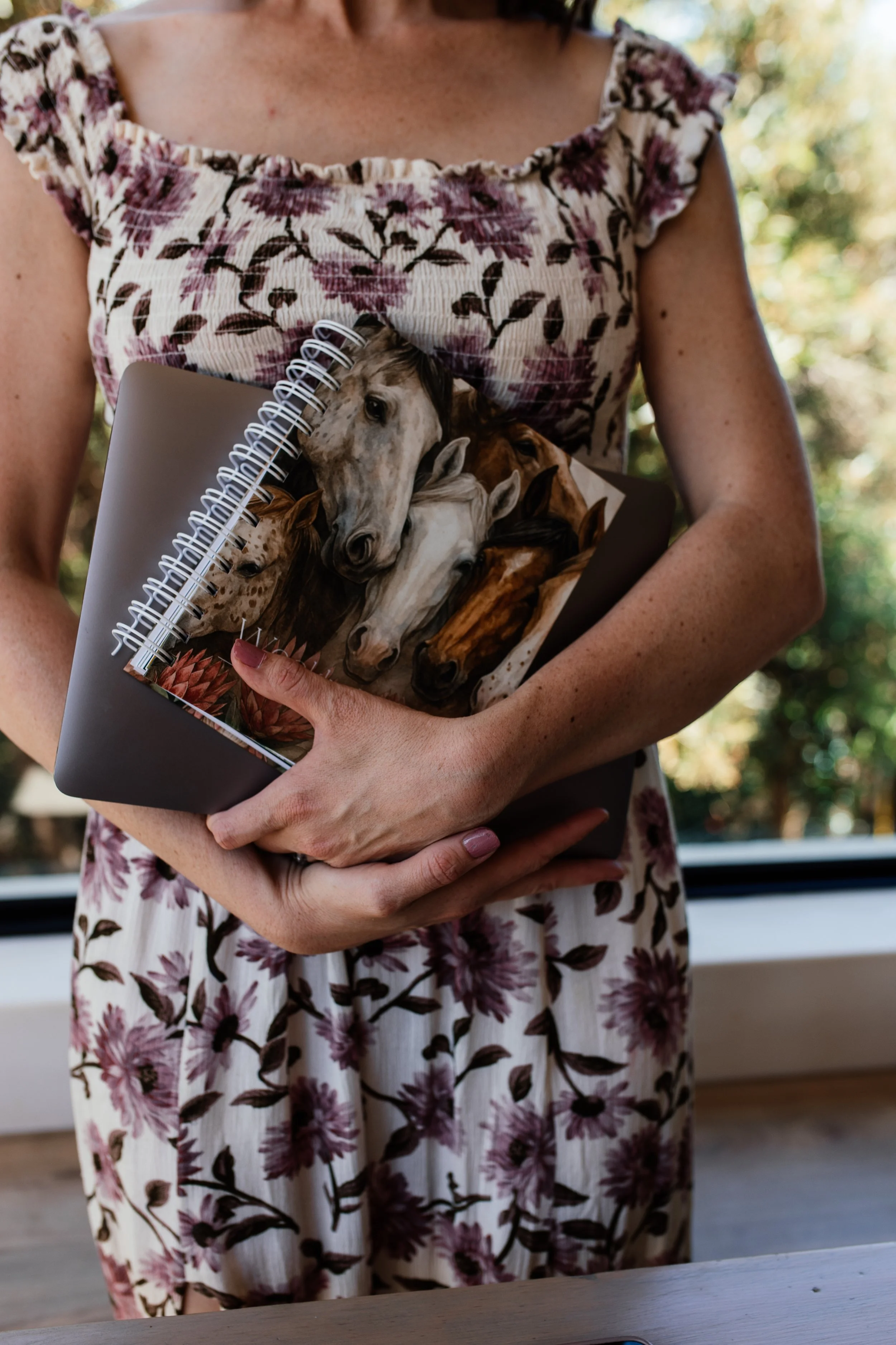 A Social media manger in a floral dress holding a notebook with horse images on the cover, standing indoors with a window and trees outside in the background.