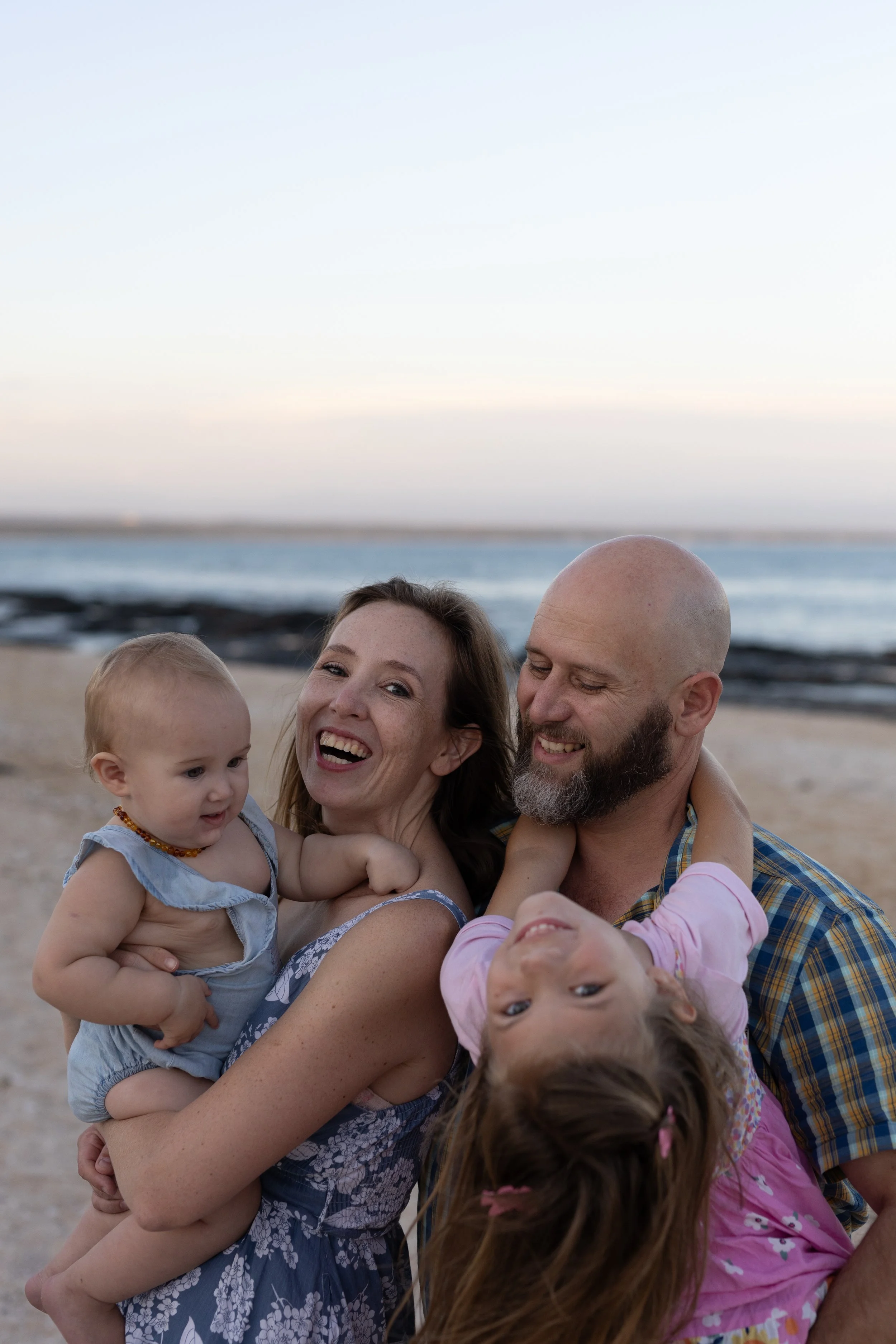 Family of four happy on the beach at sunset, with two young children, smiling faces, and ocean in the background.