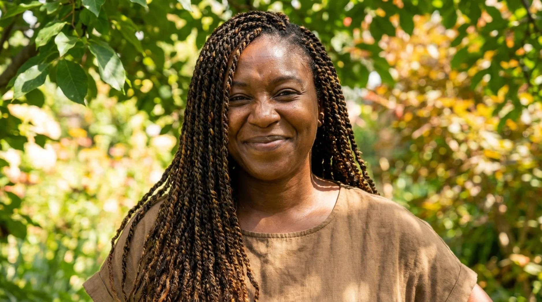 Close-up portrait of a Black woman with styled protective hair, smiling warmly outdoors in bright sunlight.