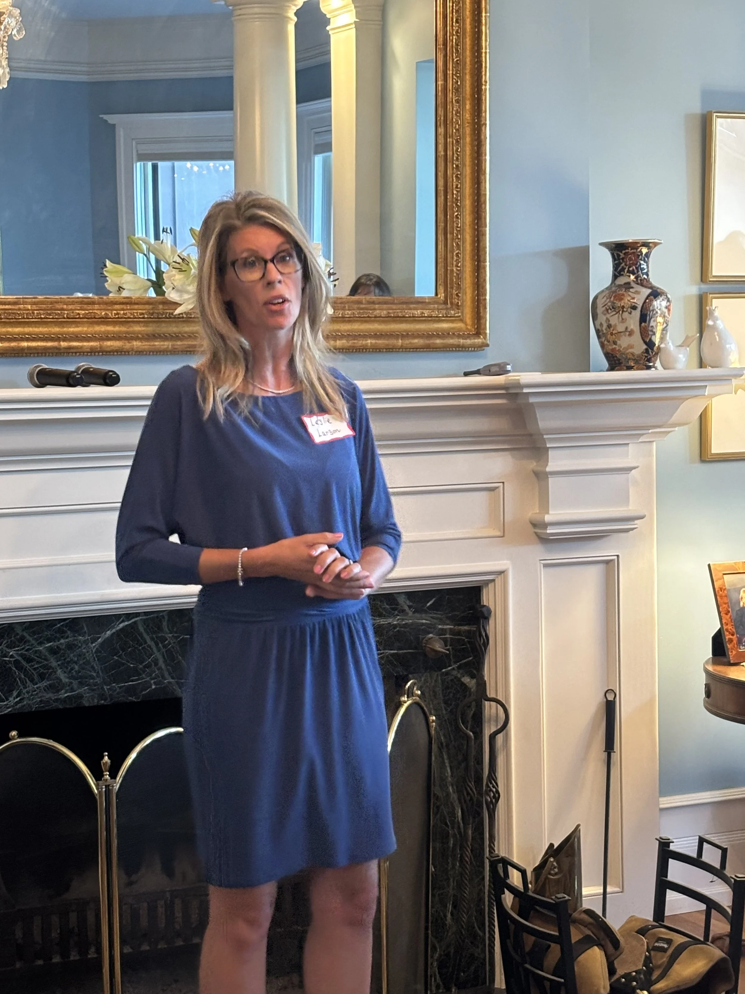 A woman with glasses and long blonde hair standing in front of a fireplace, speaking, with a large gold-framed mirror, vases, and decorative objects in the background.