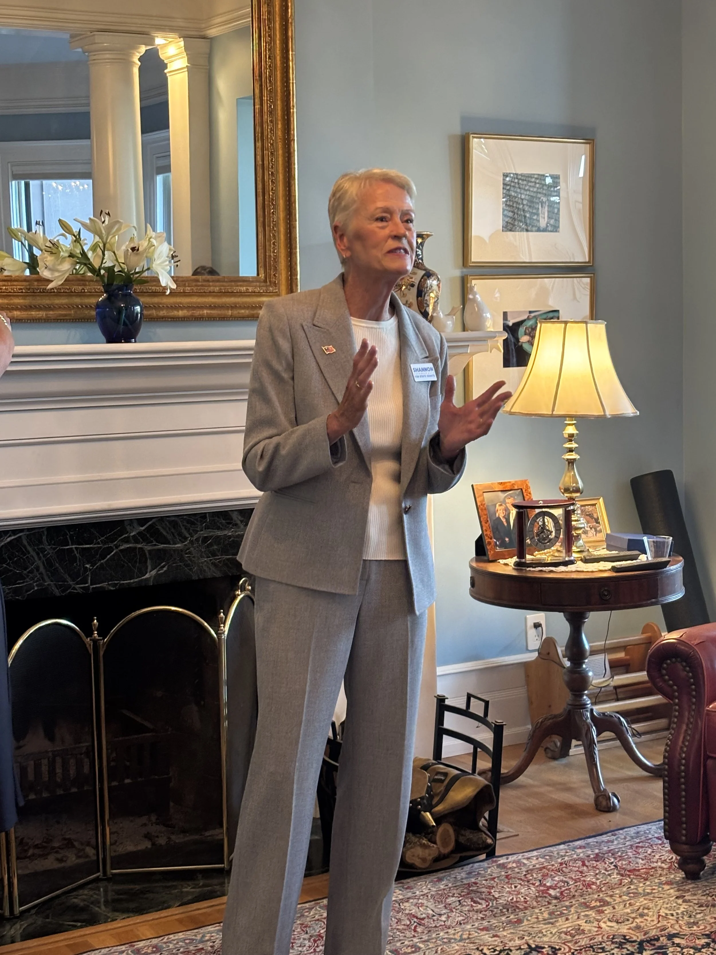 A woman with short gray hair wearing a tan blazer and white shirt standing in a living room, engaged in conversation, with a fireplace, a side table with lamps and framed photos, and decorative items behind her.