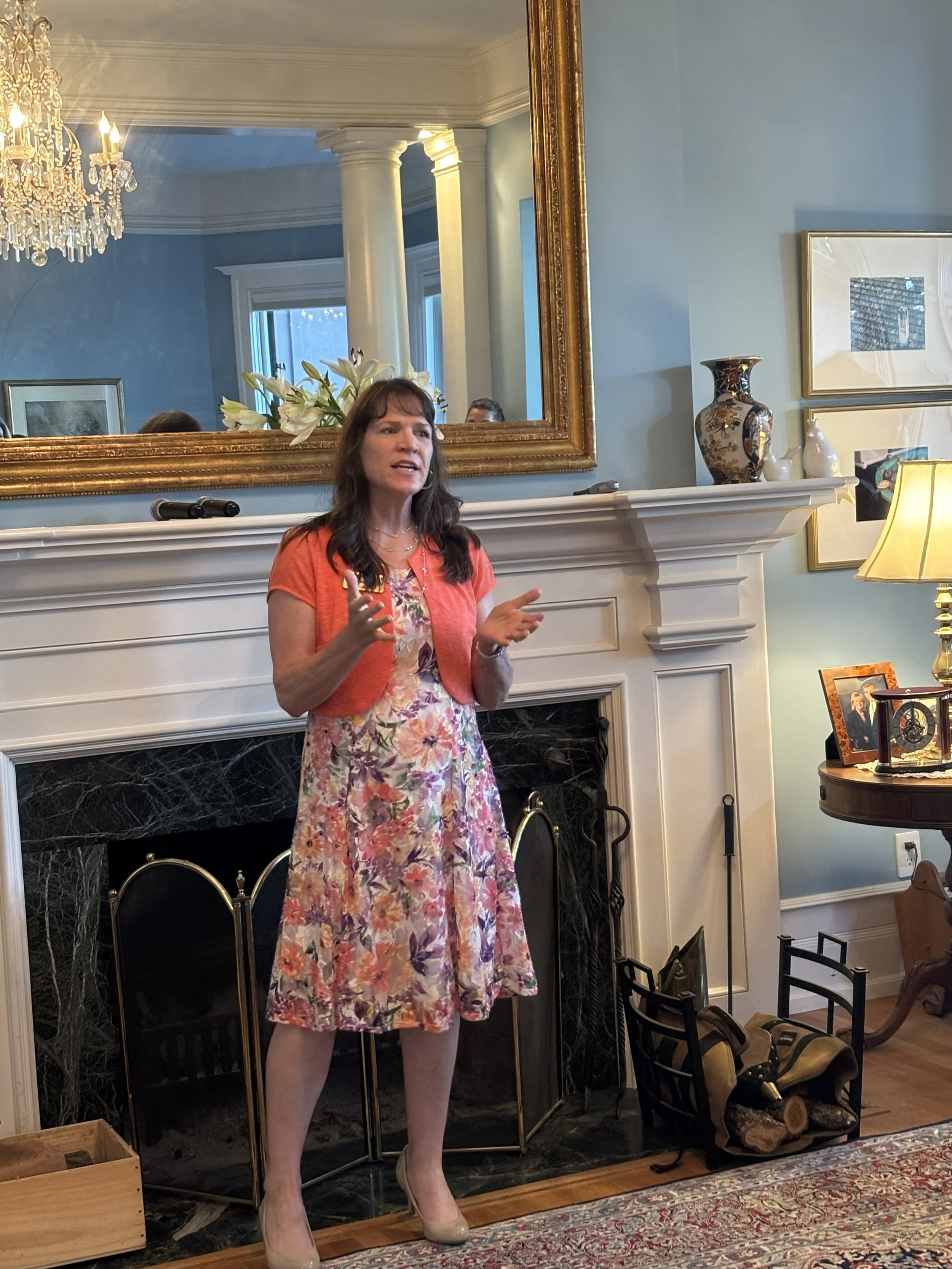 A woman with brown hair, wearing a floral dress and beige heels, speaking in a living room in front of a fireplace. The room features a large mirror, a chandelier, framed artwork, and decorative vases.