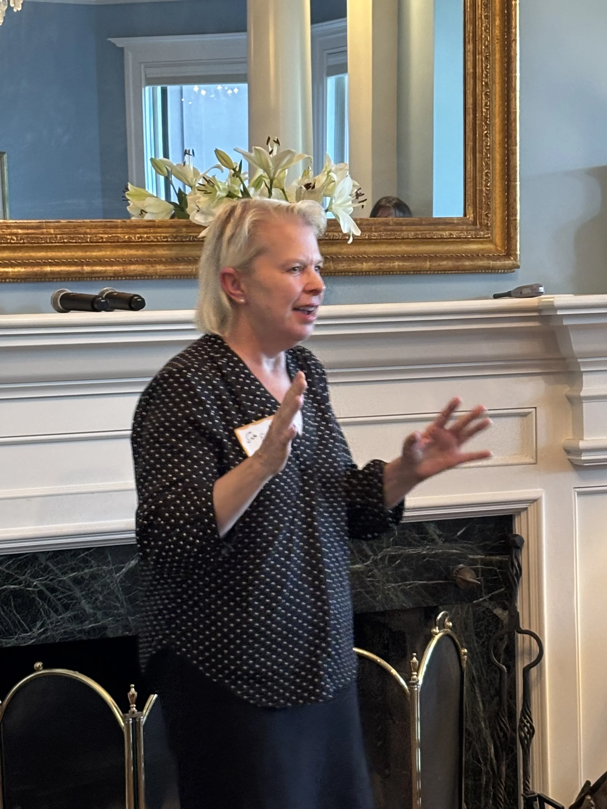 A woman with blond hair wearing a black and white polka dot top speaking in front of a mirror with a gold frame, a white flower arrangement, and two microphones on a mantelpiece in a room with blue walls.