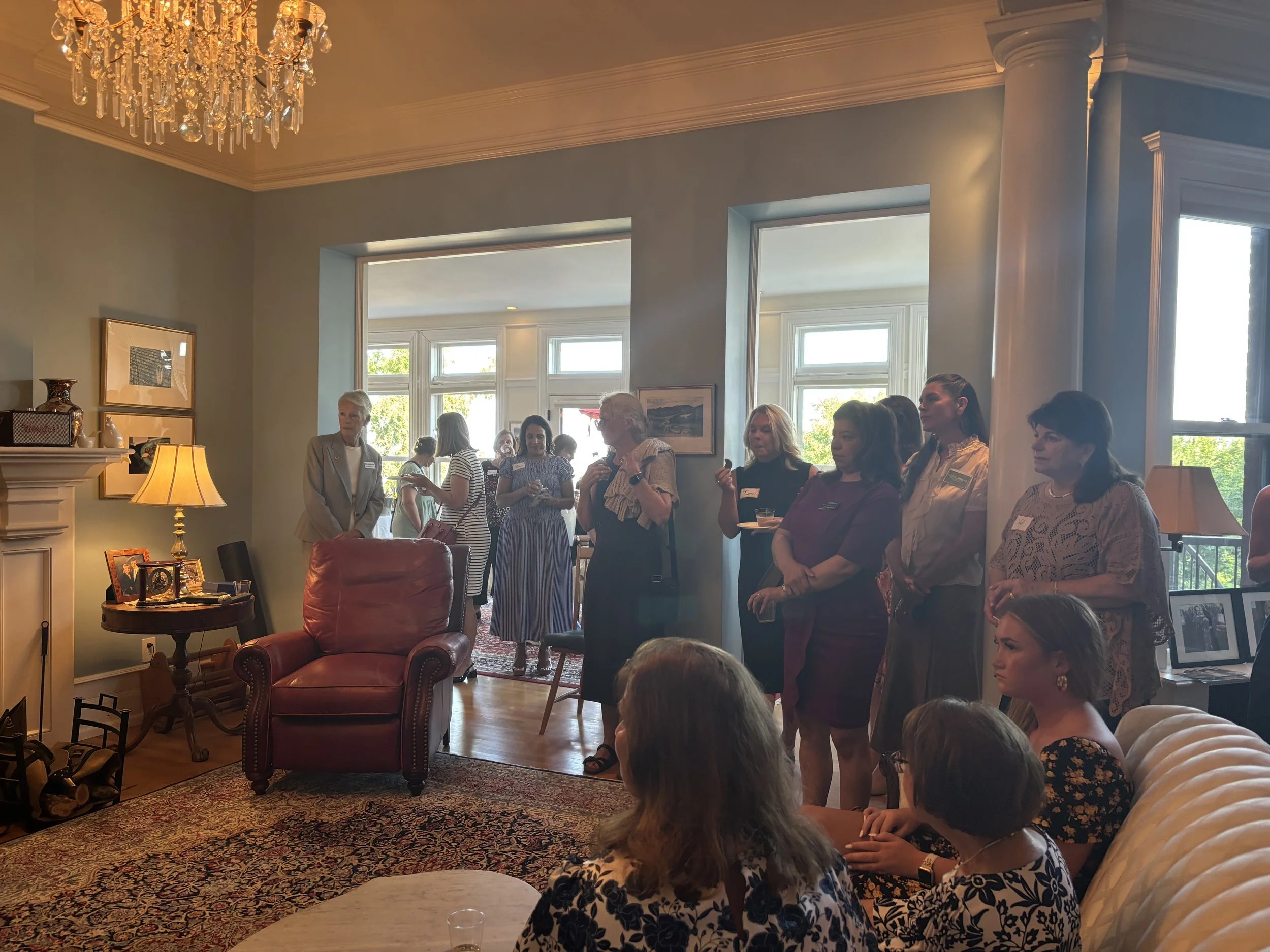 People standing and sitting in a living room during a social gathering, with a chandelier, lamps, framed photographs, and large windows showing trees outside.
