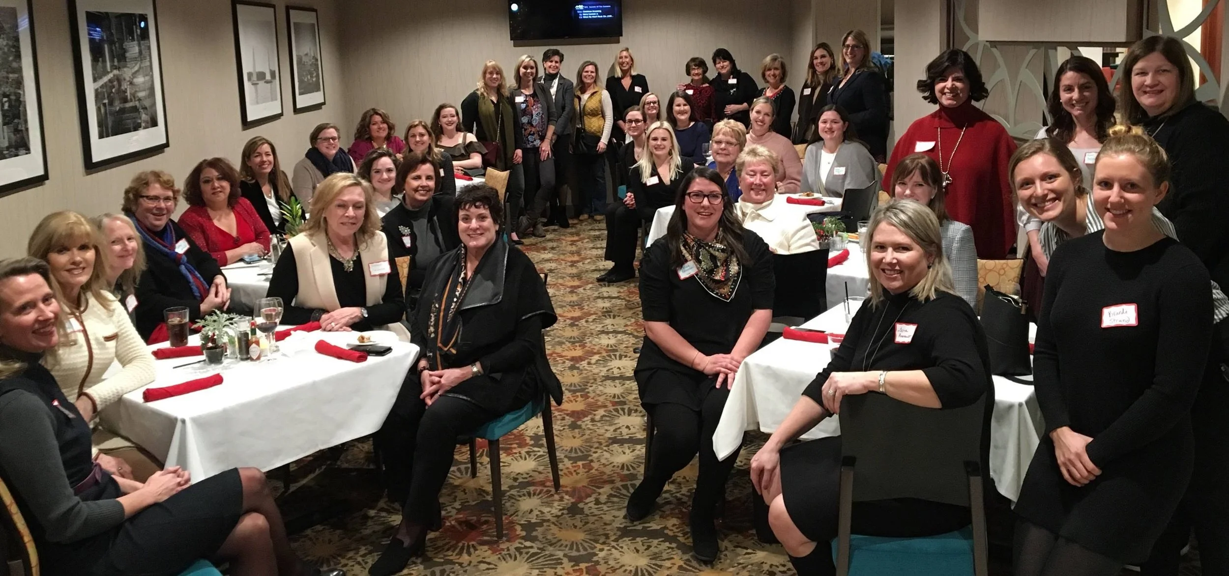 Group of women gathered at a formal event in a banquet room, some seated at tables with white tablecloths and others standing behind, smiling for the camera.