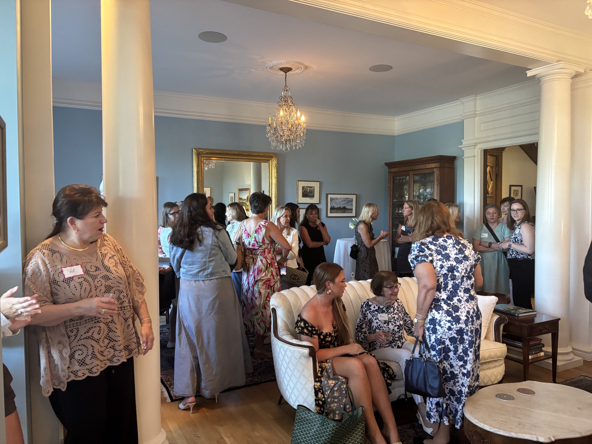 A gathering of women socializing and mingling in a well-decorated living room with classic furniture and artwork on the walls, under a chandelier lighting.