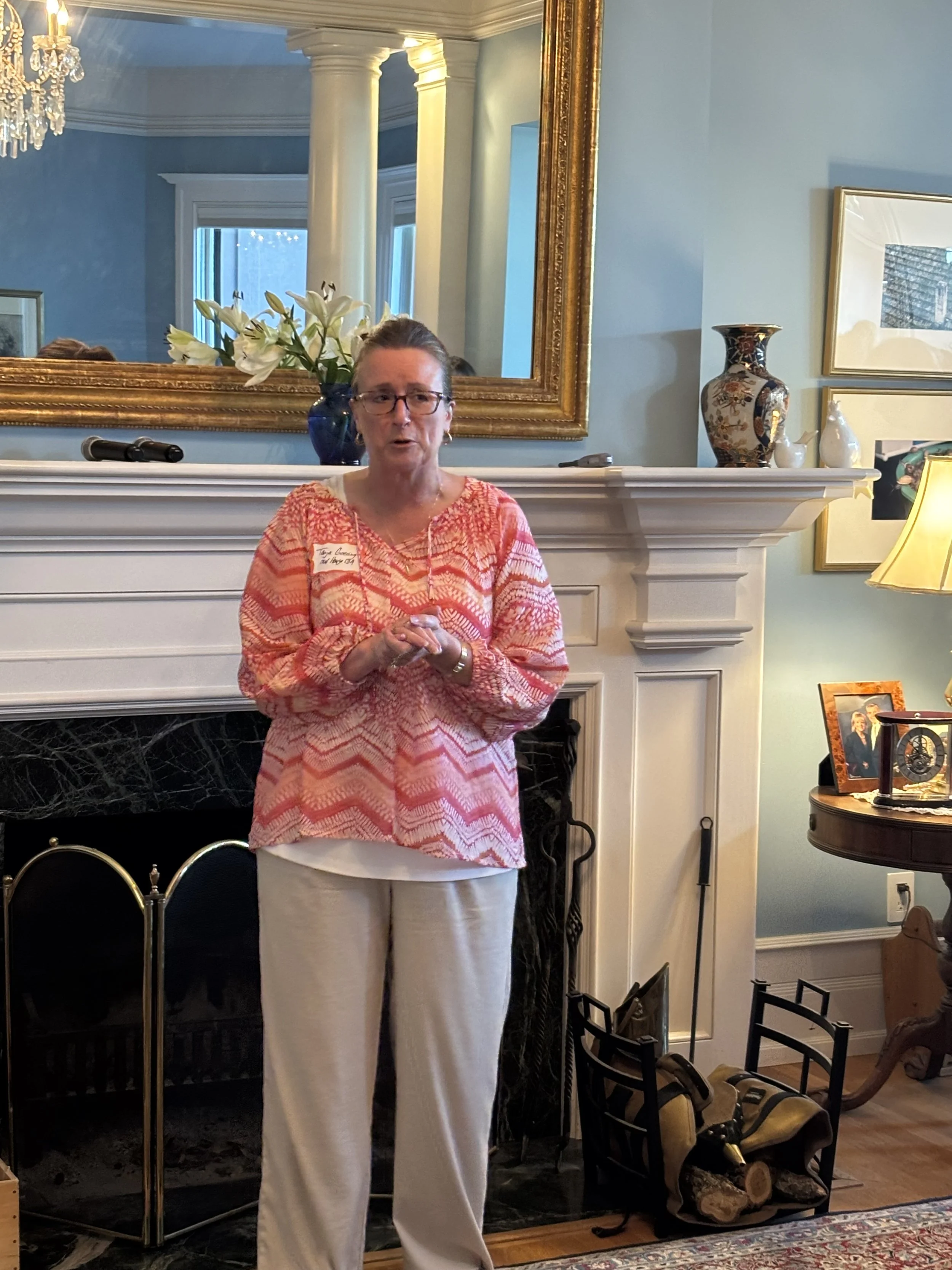 A woman standing in front of a fireplace with decorative objects and framed pictures, holding a phone, wearing glasses and a pink patterned top. A large mirror, vases, and a chandelier are visible in the background.