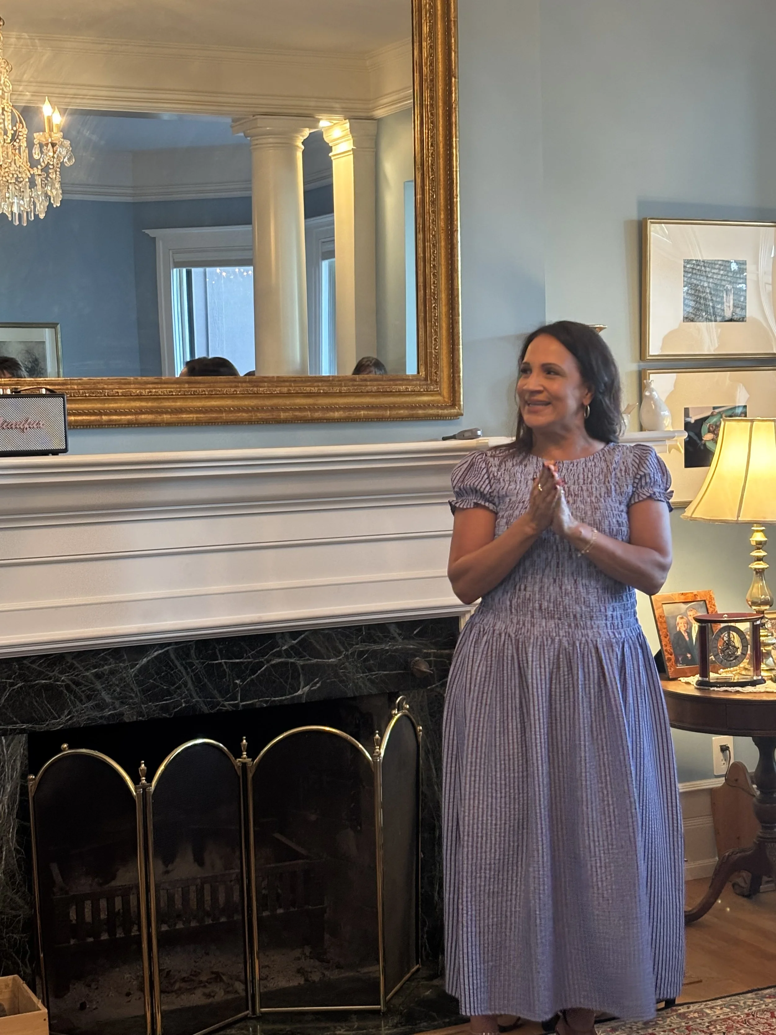 A woman in a striped dress standing in front of a fireplace, smiling and clasping her hands together, with a large gold-framed mirror and framed pictures on the wall behind her, a table with a lamp to her right.