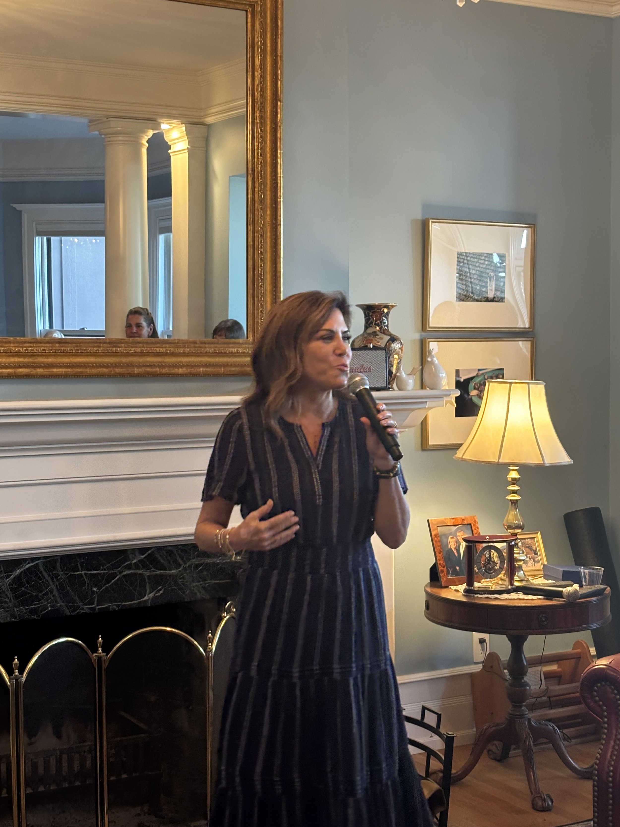 A woman with light brown hair holding a microphone in a living room, with framed photographs, decorative vases, a lamp, and furniture in the background. A large mirror on the wall reflects a woman and two children.