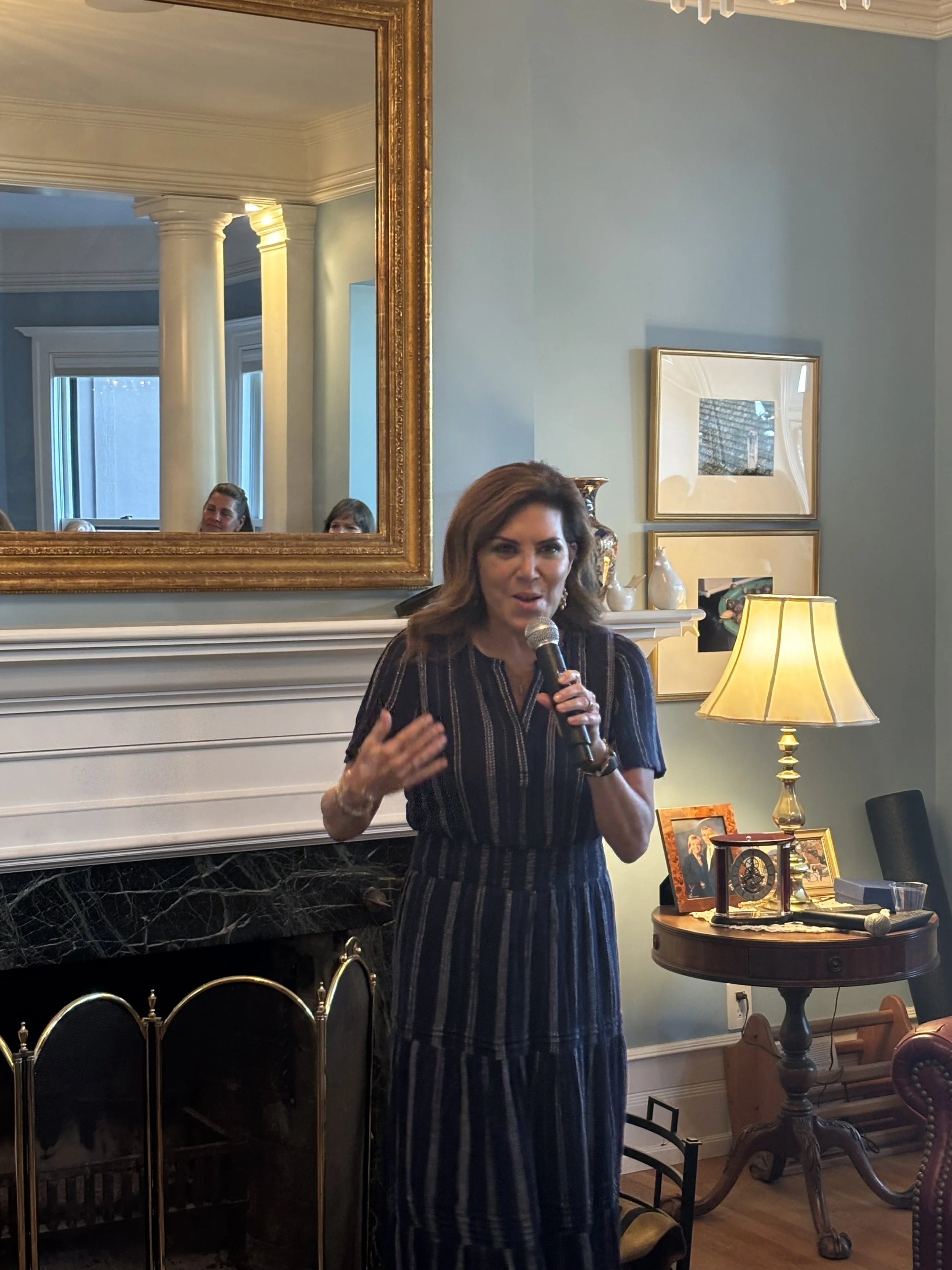 Woman with brown hair speaking into a microphone in a living room with a large gold-framed mirror, a fireplace, a side table with framed photos, a lamp, and decorative items.