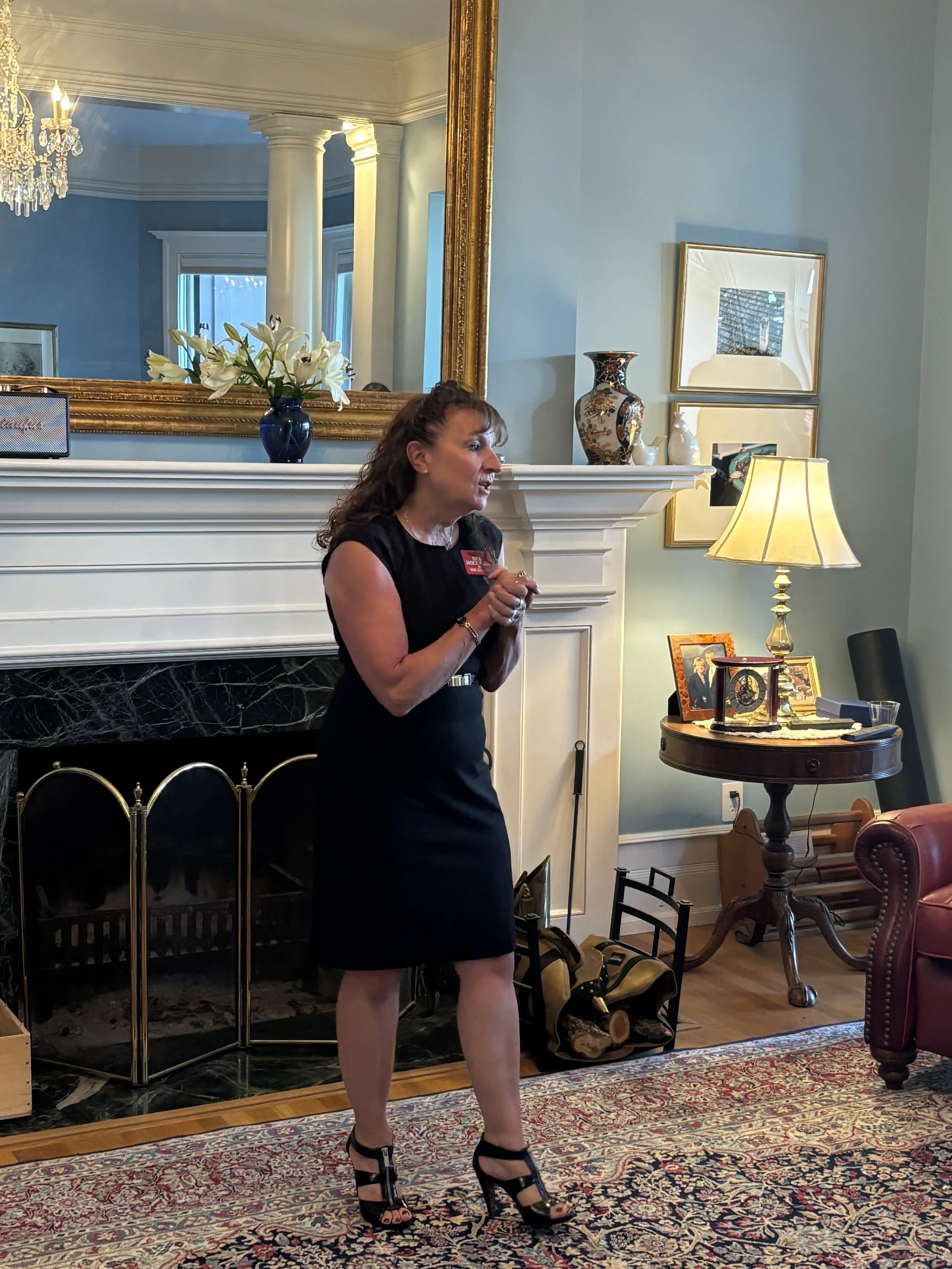 A woman in a black cocktail dress and black high heels stands near a fireplace with a large mirror, decorative vases, and framed photos on the mantel. The room has blue walls, a chandelier, and a patterned rug on the floor.