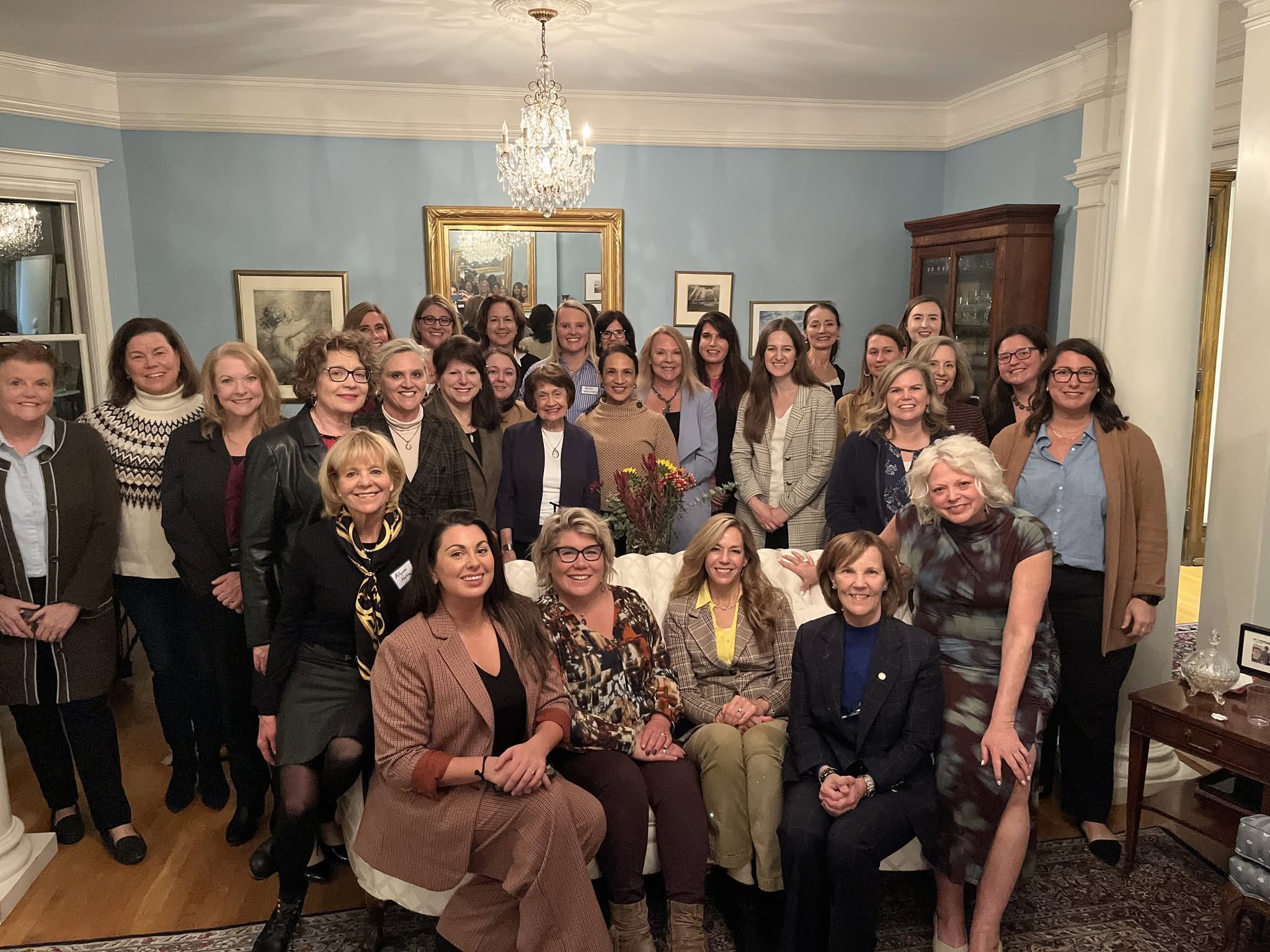 Group of women gathered in a decorated living room, smiling for a group photo in front of a wall with artwork and a chandelier hanging from the ceiling.