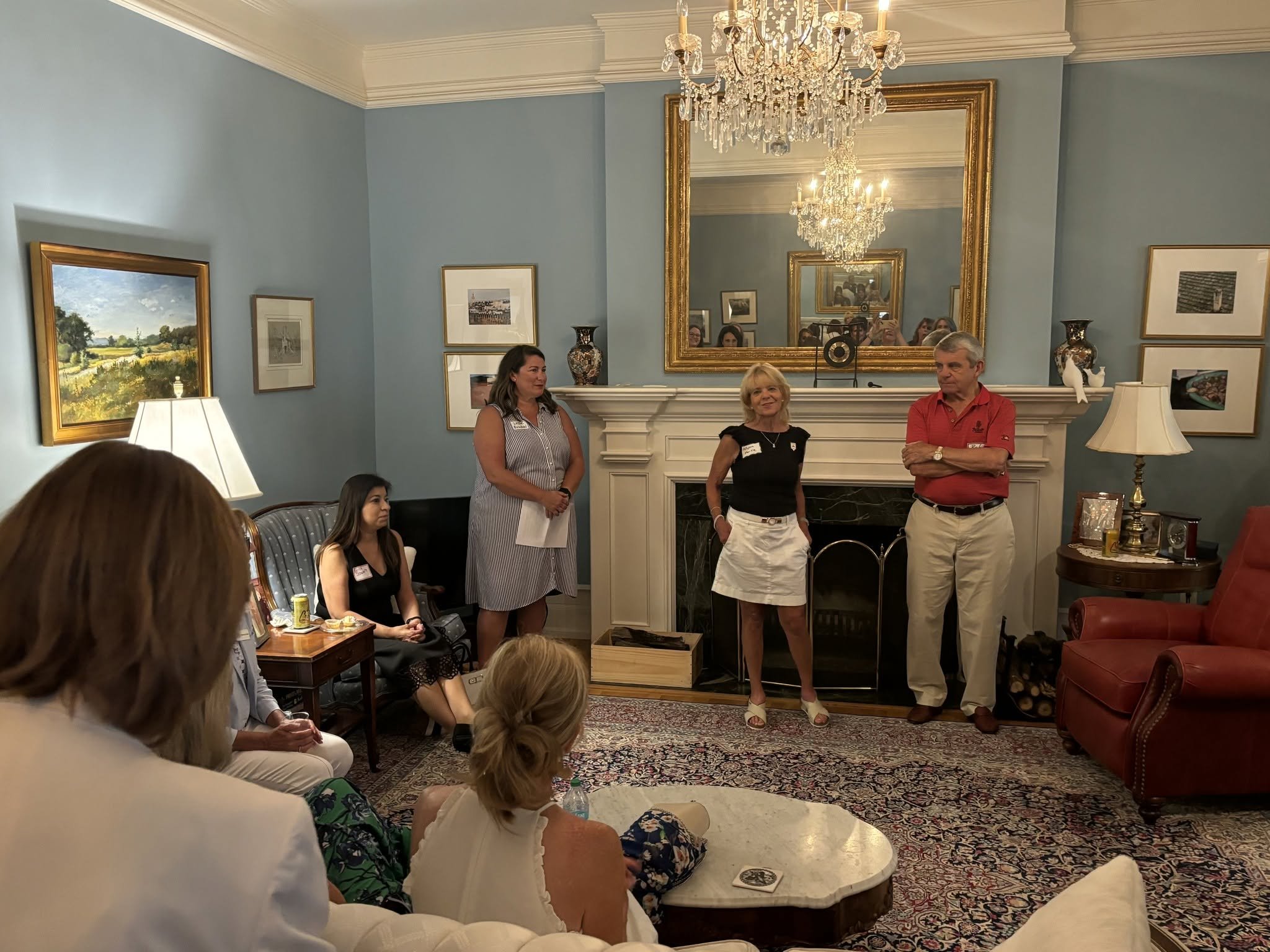 People gathered in a living room with a fireplace, blue walls, and artwork, listening to a woman speak. A large mirror above the fireplace reflects a chandelier and additional attendees, with some individuals sitting and others standing.