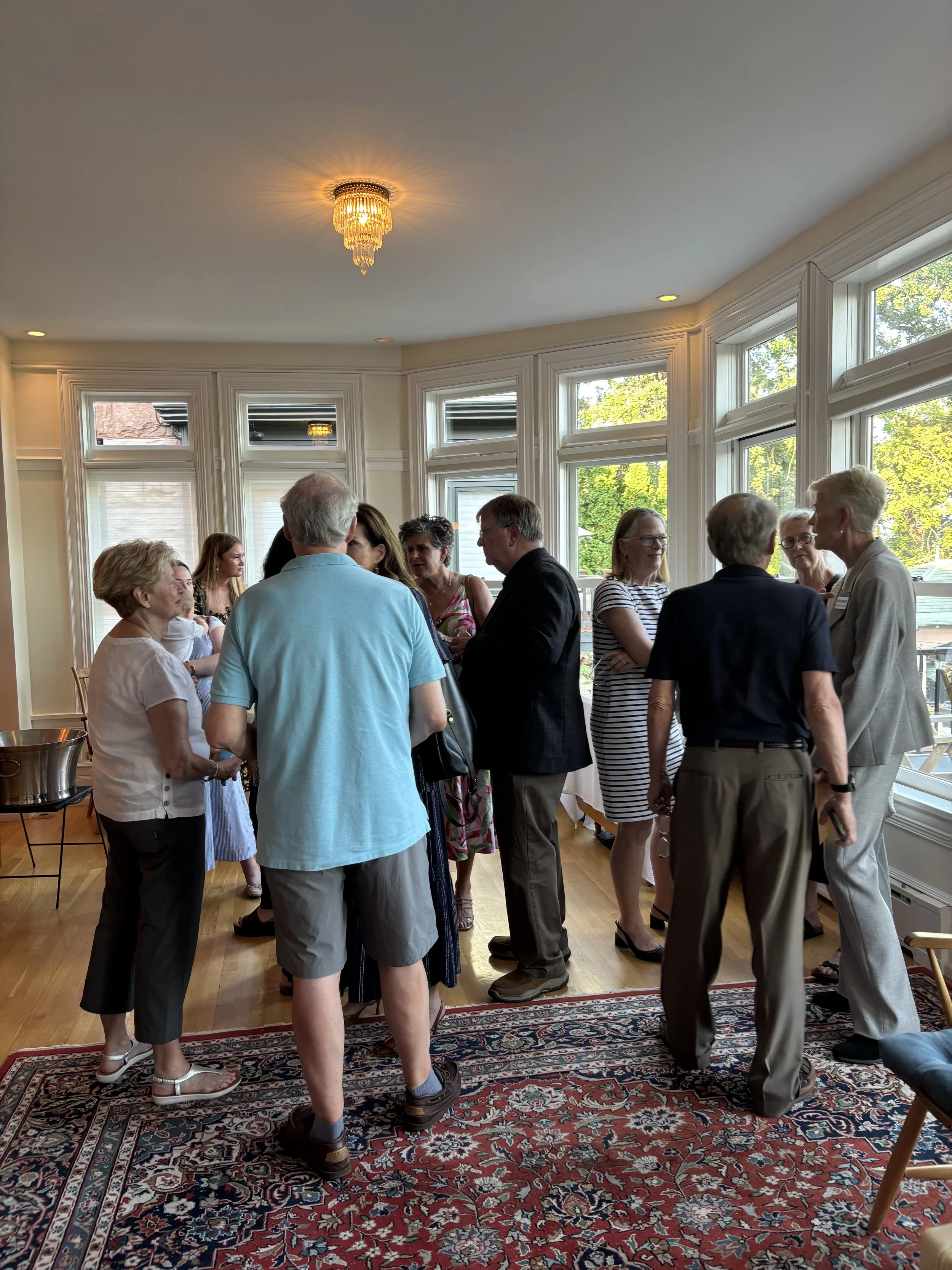 A group of people, mostly middle-aged and elderly, gathered in a bright room with large windows, engaging in conversation and socializing, with some smiling and talking in pairs, during daytime.