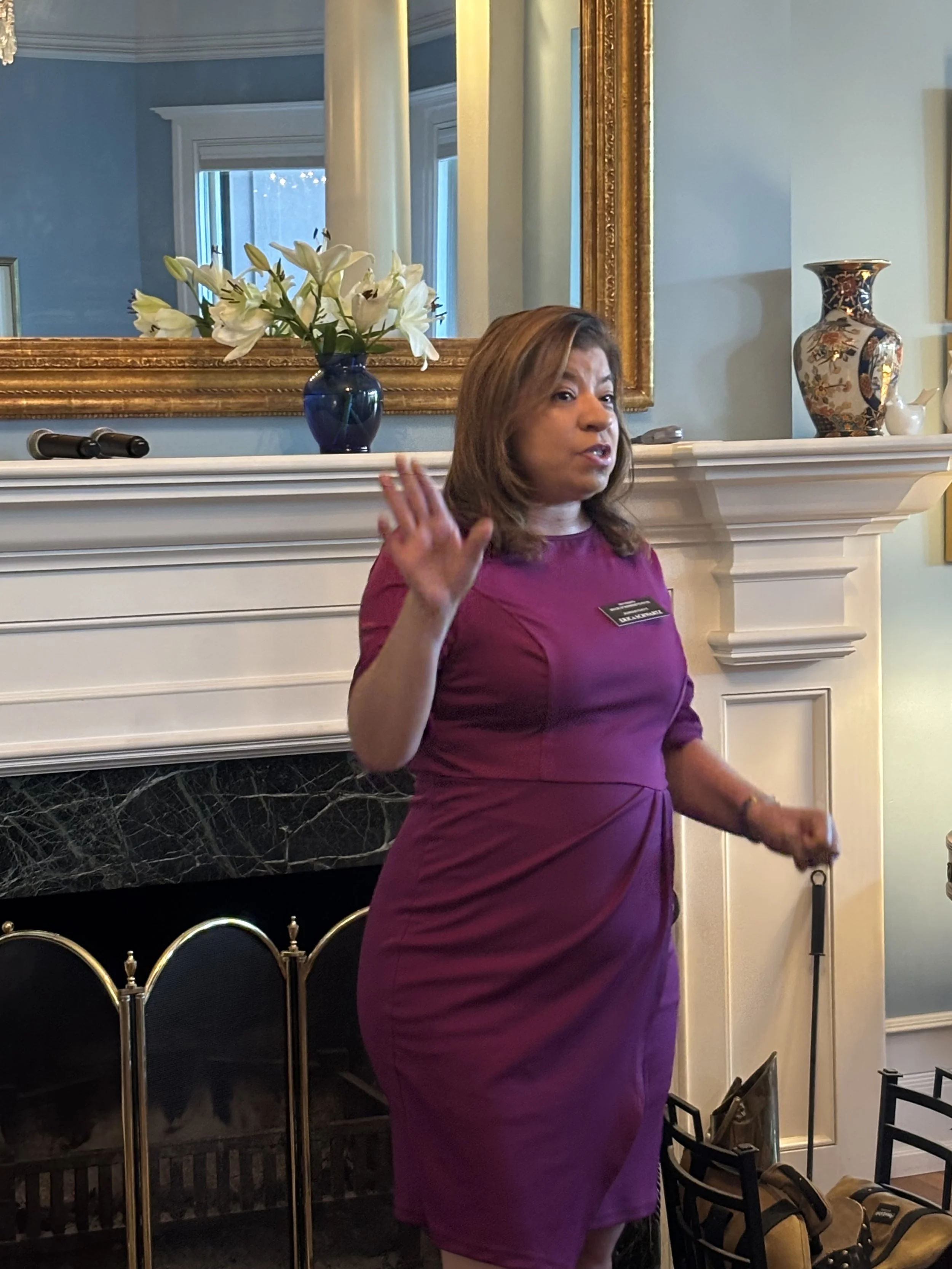 A woman in a purple dress gesturing with her right hand, standing in front of a fireplace with flowers and decorative vases, with chairs and bags on the floor nearby.