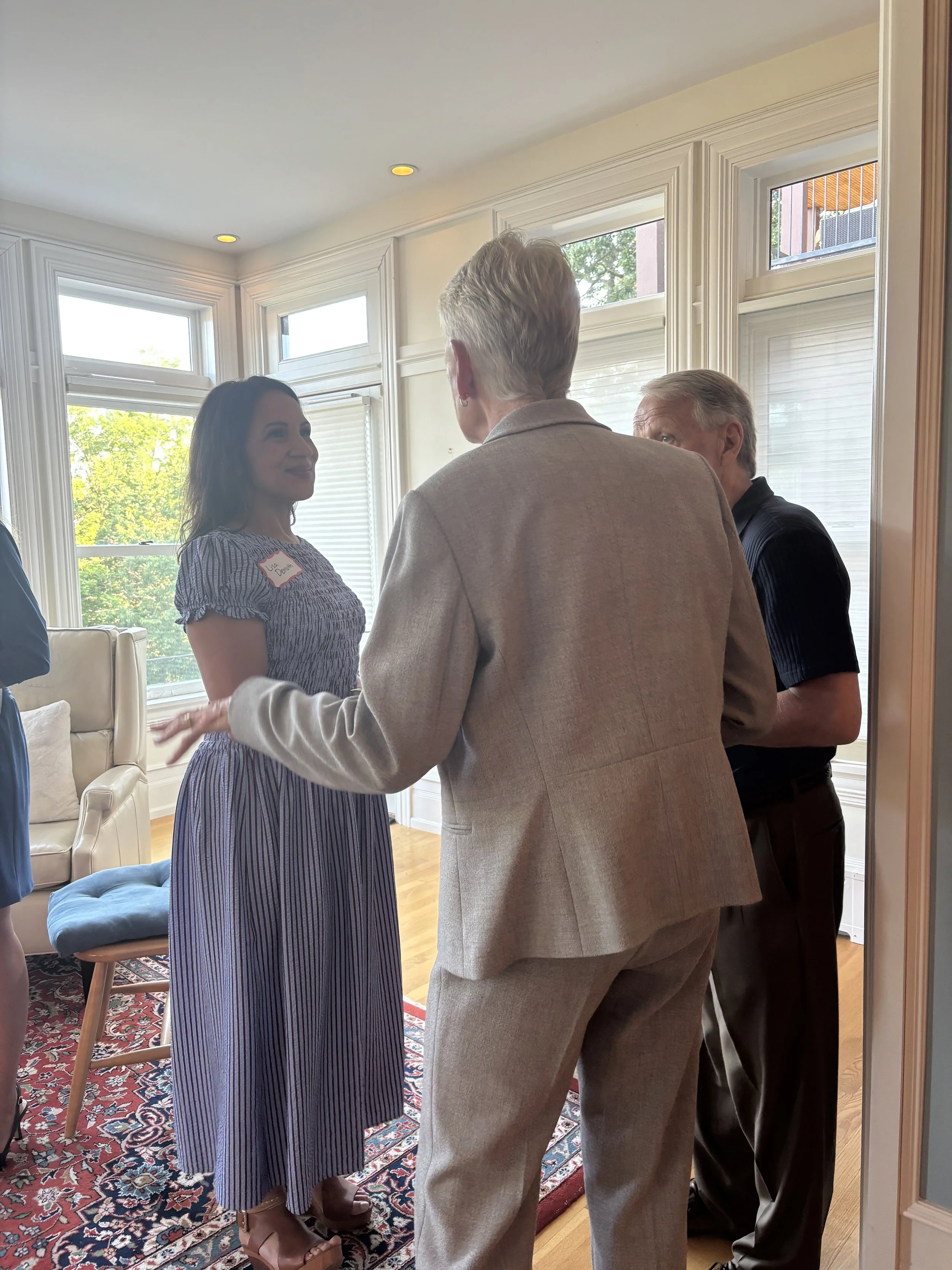 Four people are engaged in conversation inside a bright room with large windows. One woman with dark hair, wearing a striped dress and a name tag, stands facing three men. One man with gray hair and a beige blazer faces away from the camera, another 