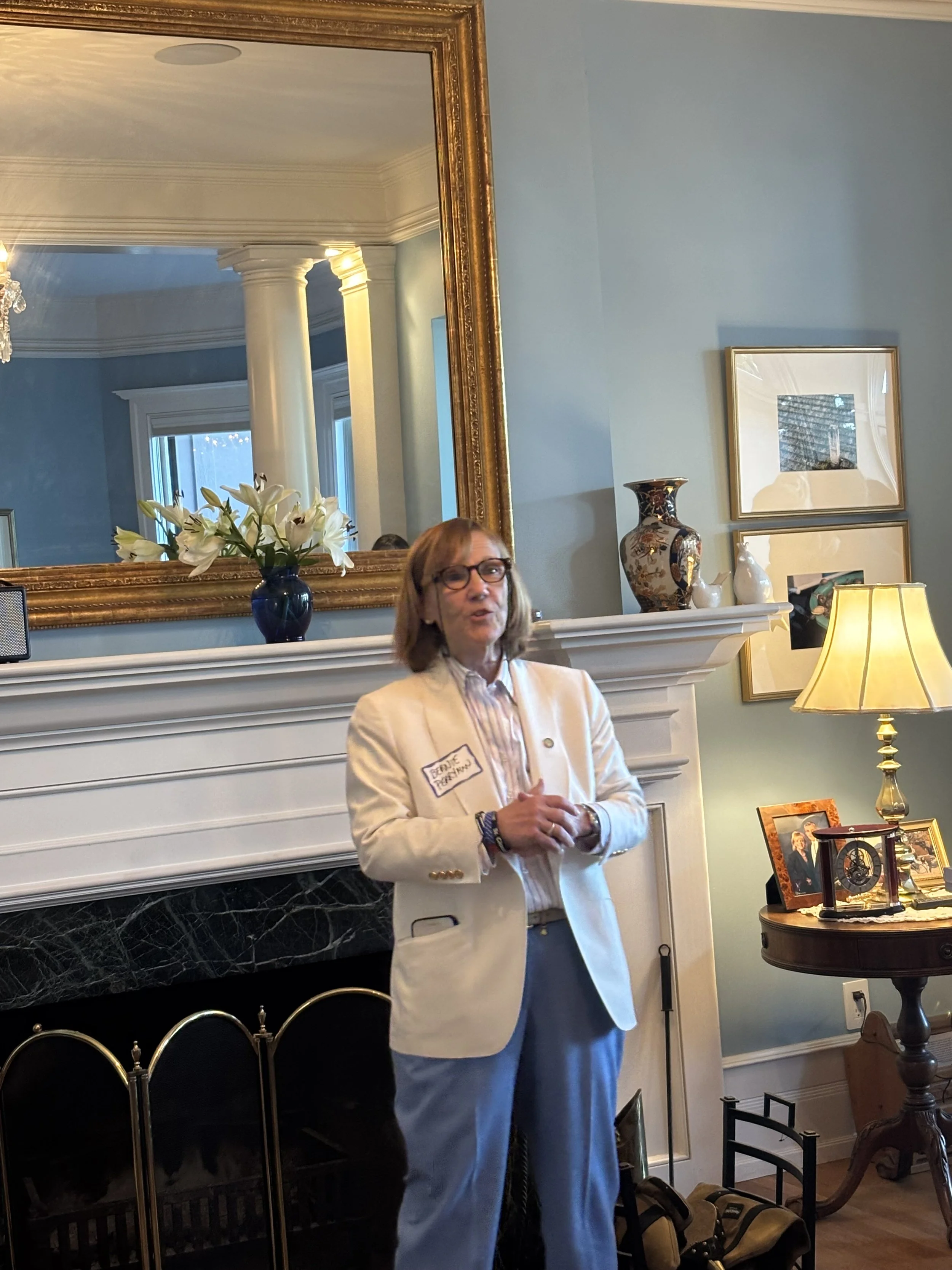 A woman wearing glasses and a white blazer stands in front of a fireplace, speaking. The fireplace has a black marble surround and a white mantel with framed photographs, a decorative clock, and a small lamp. Behind her is a large gold-framed mirror,