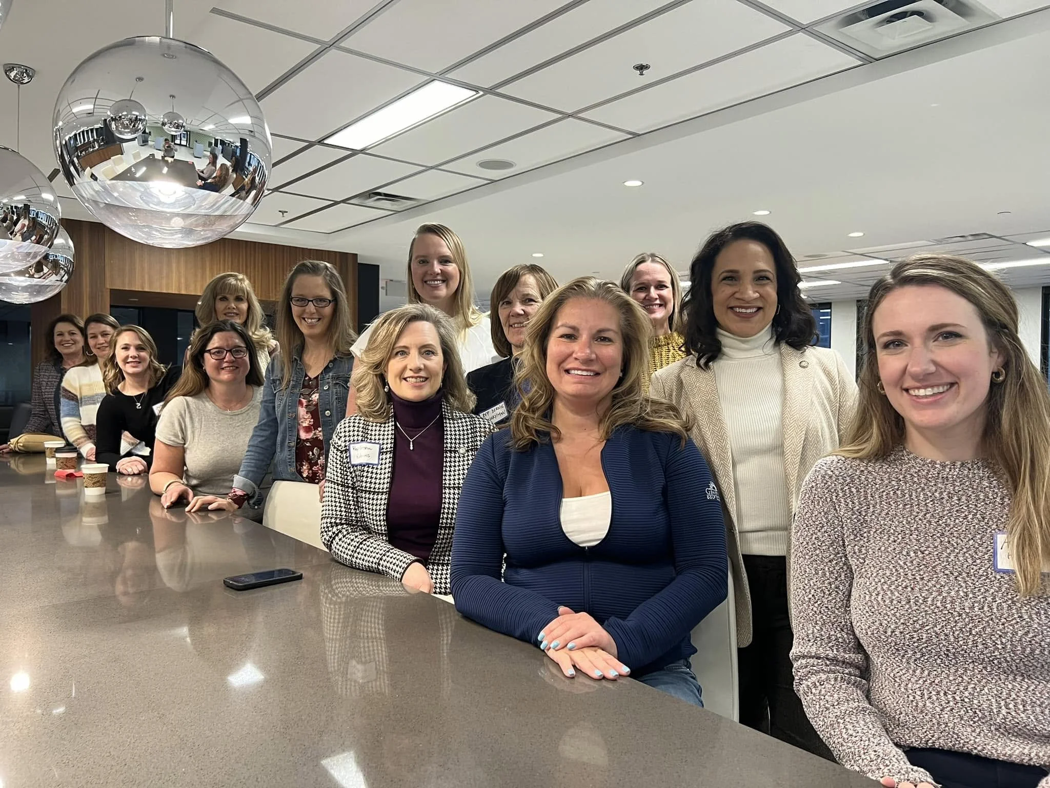 Group of women gathered around a table in an office or conference room, smiling at the camera, with a reflective spherical light fixture hanging from the ceiling.