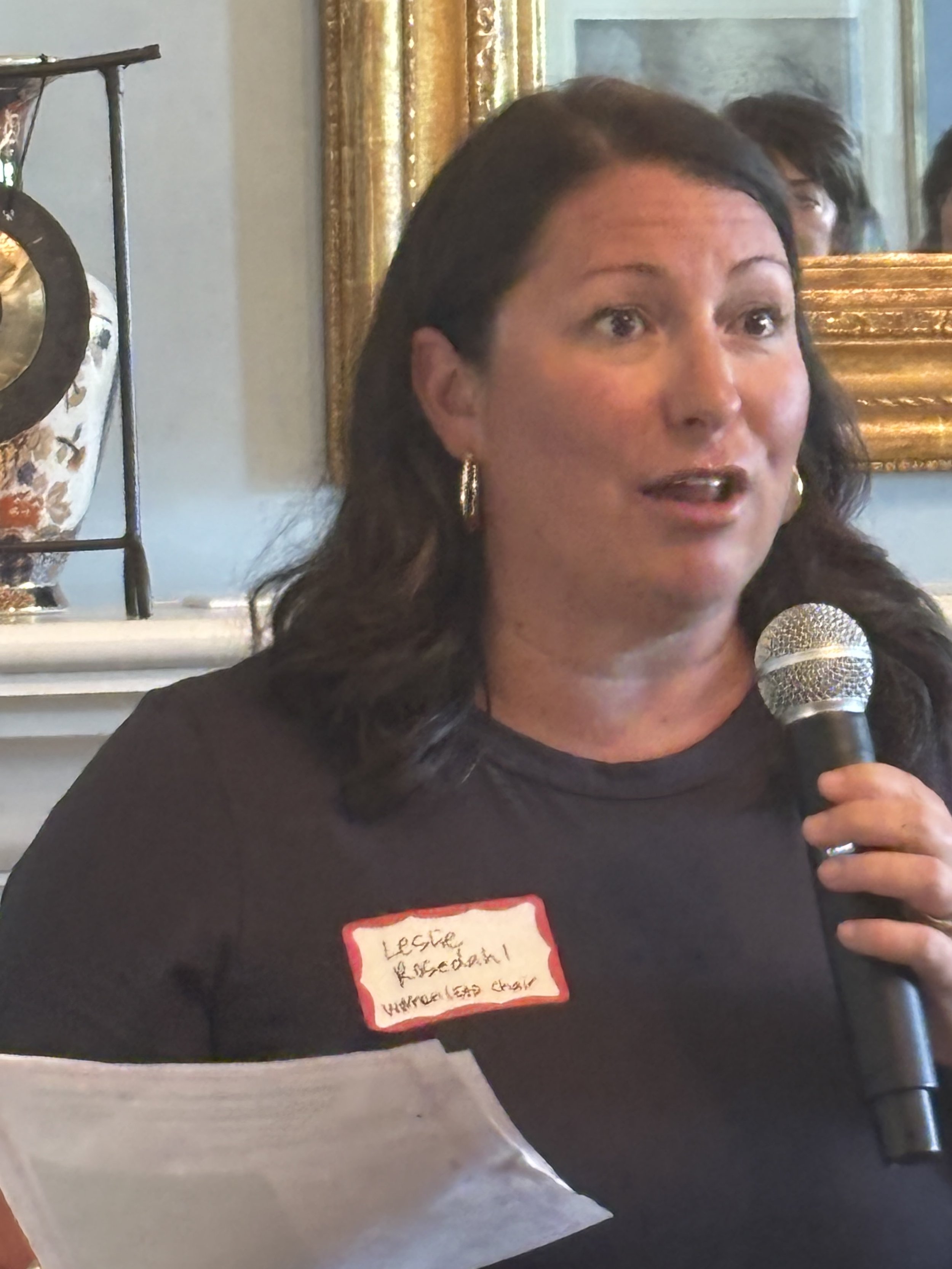 A woman with long dark hair holding a microphone speaks at an event. She wears a black shirt and has a name tag that reads "Leslie Kessaiyah!". In the background, there is a framed mirror and a decorative object with a stand, with gold-colored curtai